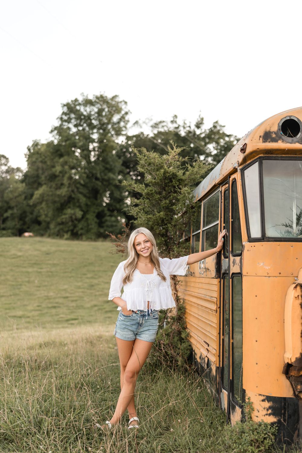 Woman in white top and denim shorts poses beside vintage yellow school bus in grassy rural setting during summer.
