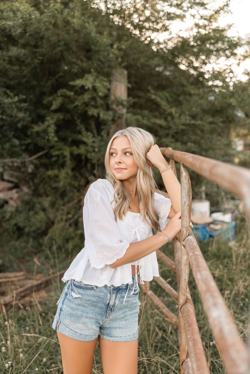 A person in a white blouse and denim shorts leans against a rustic wooden fence in a natural outdoor setting at sunset.