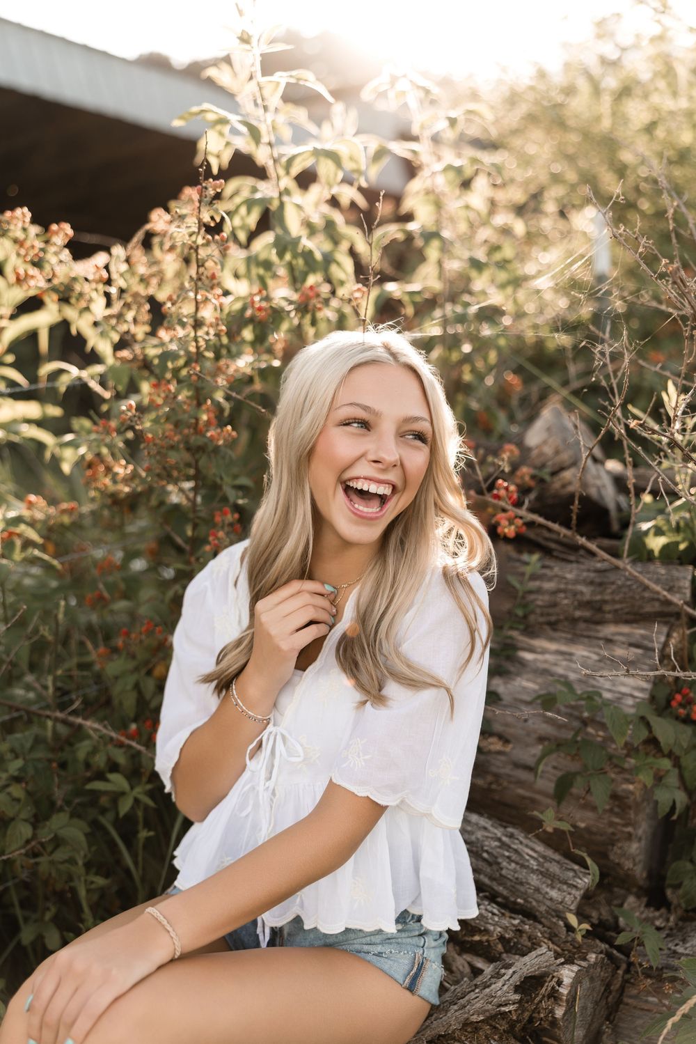 Candid outdoor portrait in white peasant blouse and denim shorts sitting by rustic wood against sunlit foliage.