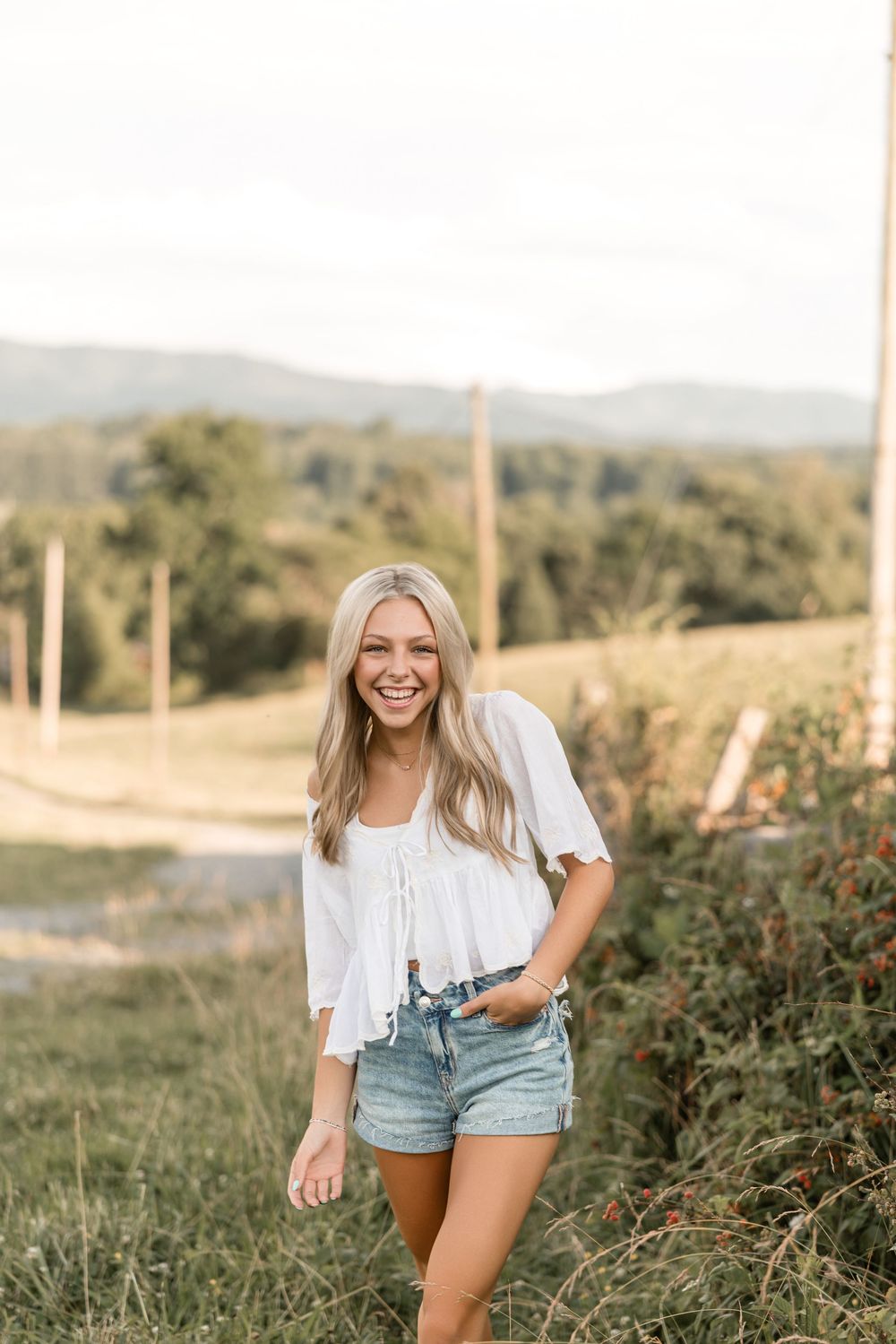 Person in a white top and denim shorts stands on a grassy hillside path with mountains in the background.