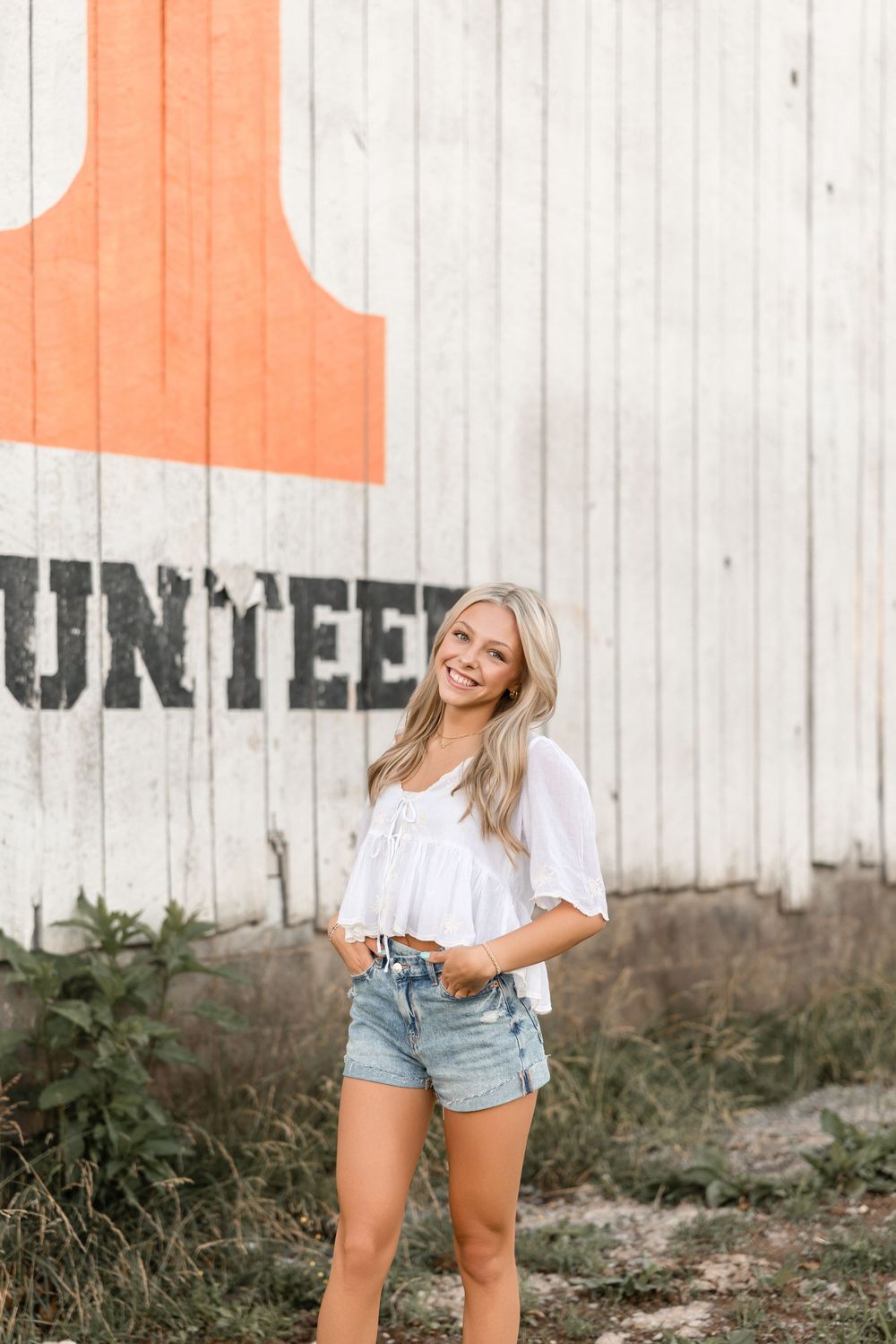 Young woman in white t-shirt and denim shorts laughs while standing against white and orange painted wall outdoors.