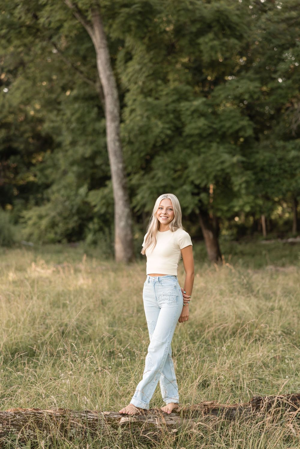 A person in a white t-shirt and light jeans stands in a grassy field with trees in the background.