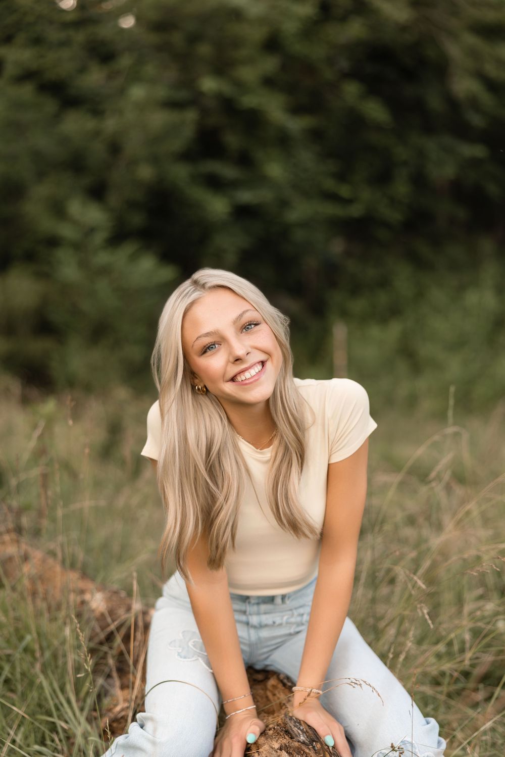 Young woman in light top and jeans sits in grass field while laughing and smiling during outdoor photo session.
