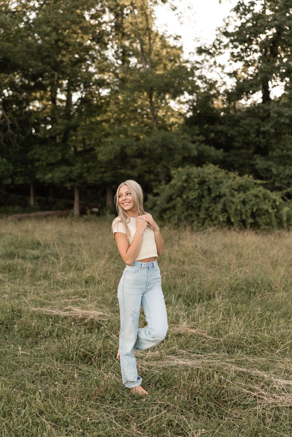 A person stands in a sunlit meadow wearing light blue jeans and a white top with trees in the background.