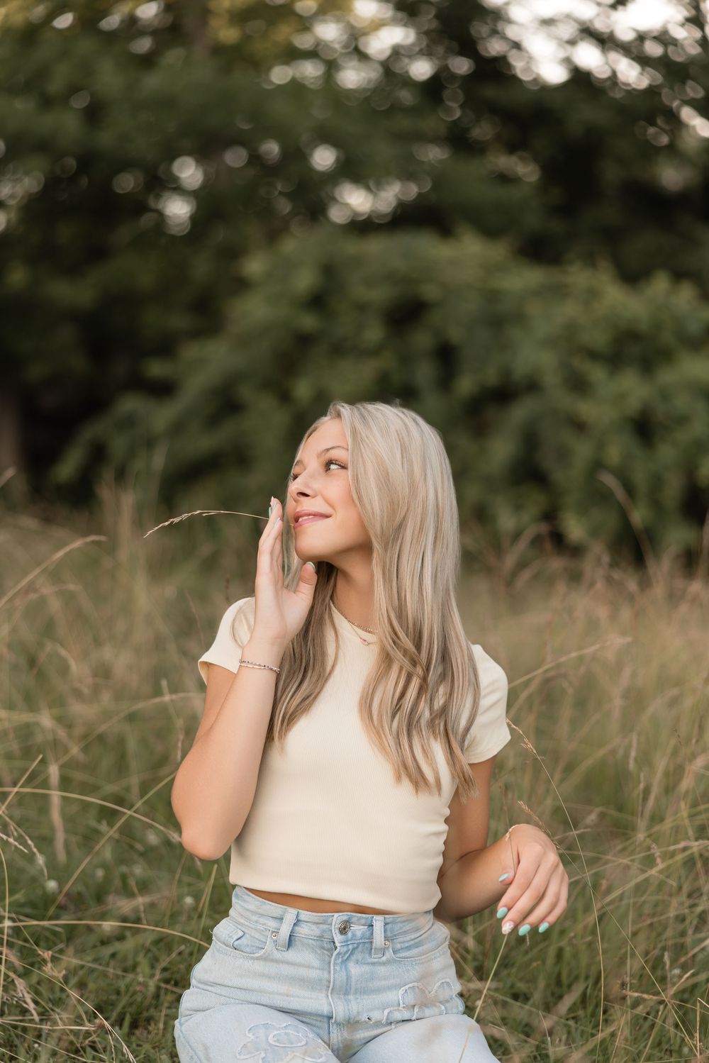 Person in a white top and light denim jeans laughing while sitting in tall grass on a sunny day.