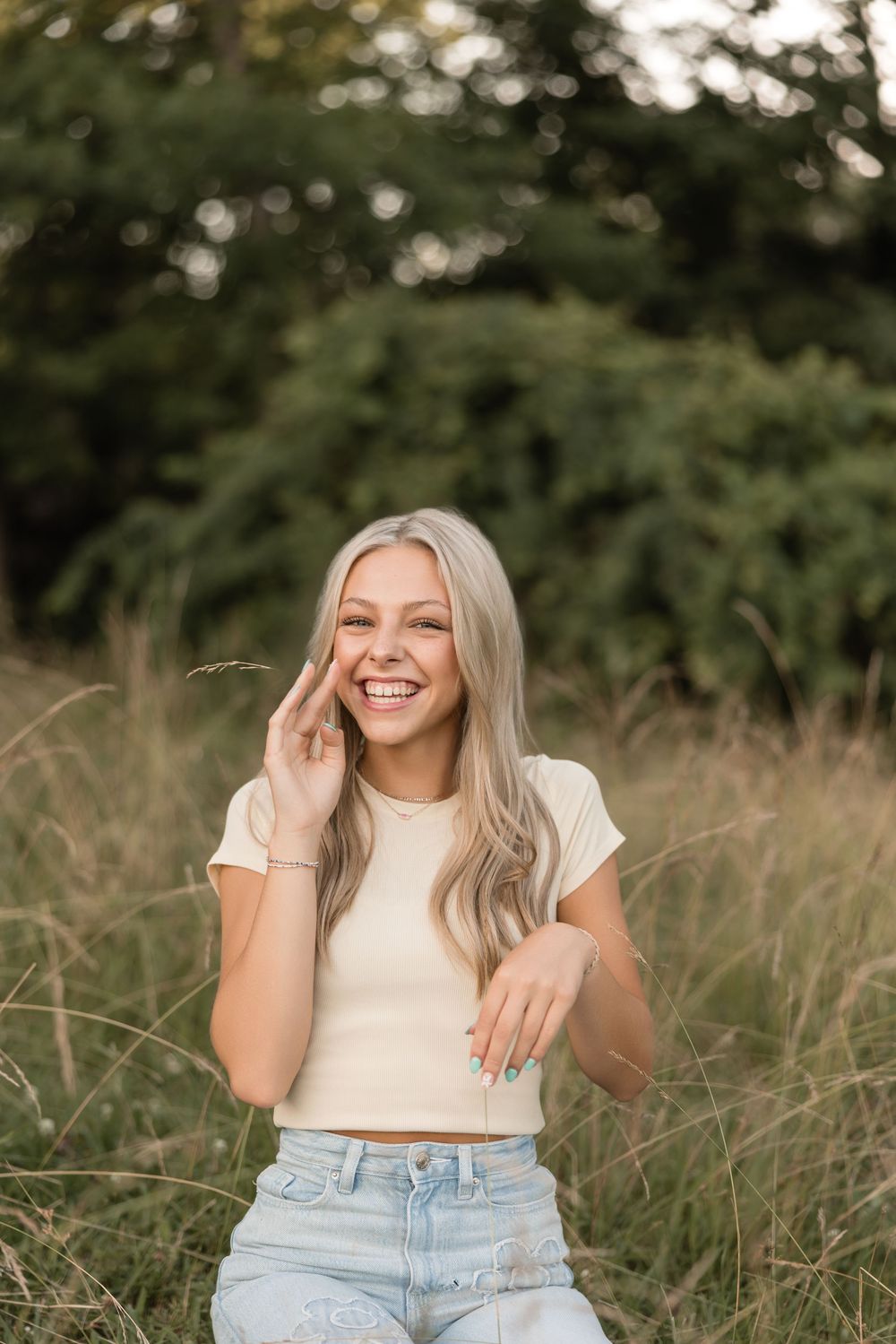 Person in white crop top and light denim jeans poses outdoors in tall grass during golden hour photography session.