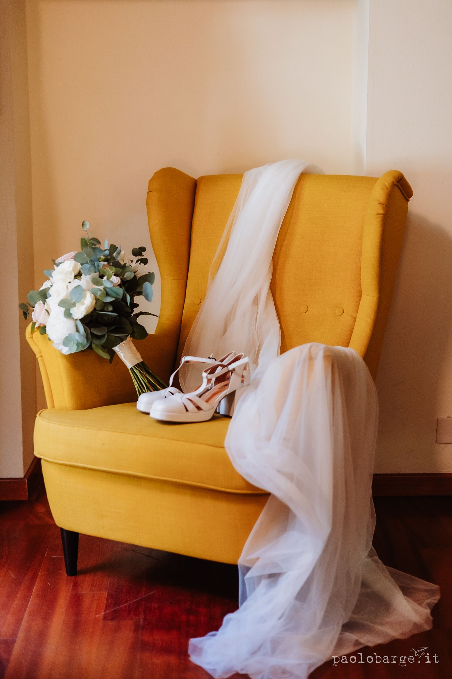 Wedding accessories including a white floral bouquet, bridal veil and strappy heels arranged on a mustard yellow armchair.