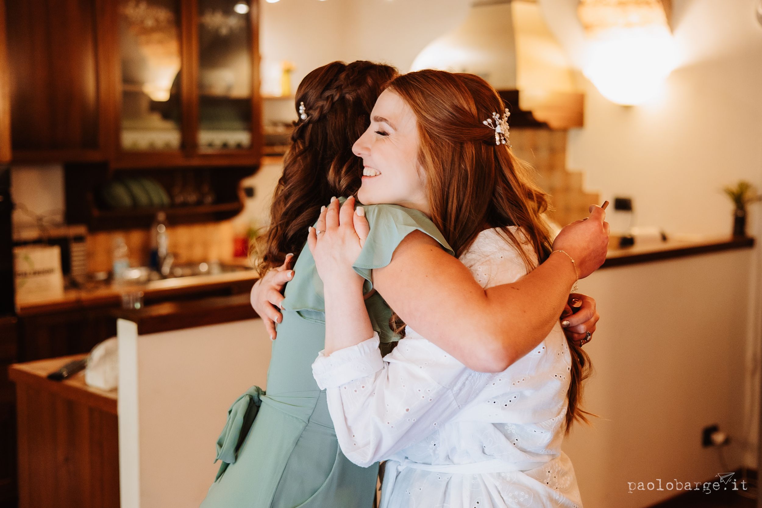 Two people in white and green dresses embrace warmly in a warmly lit kitchen setting.
