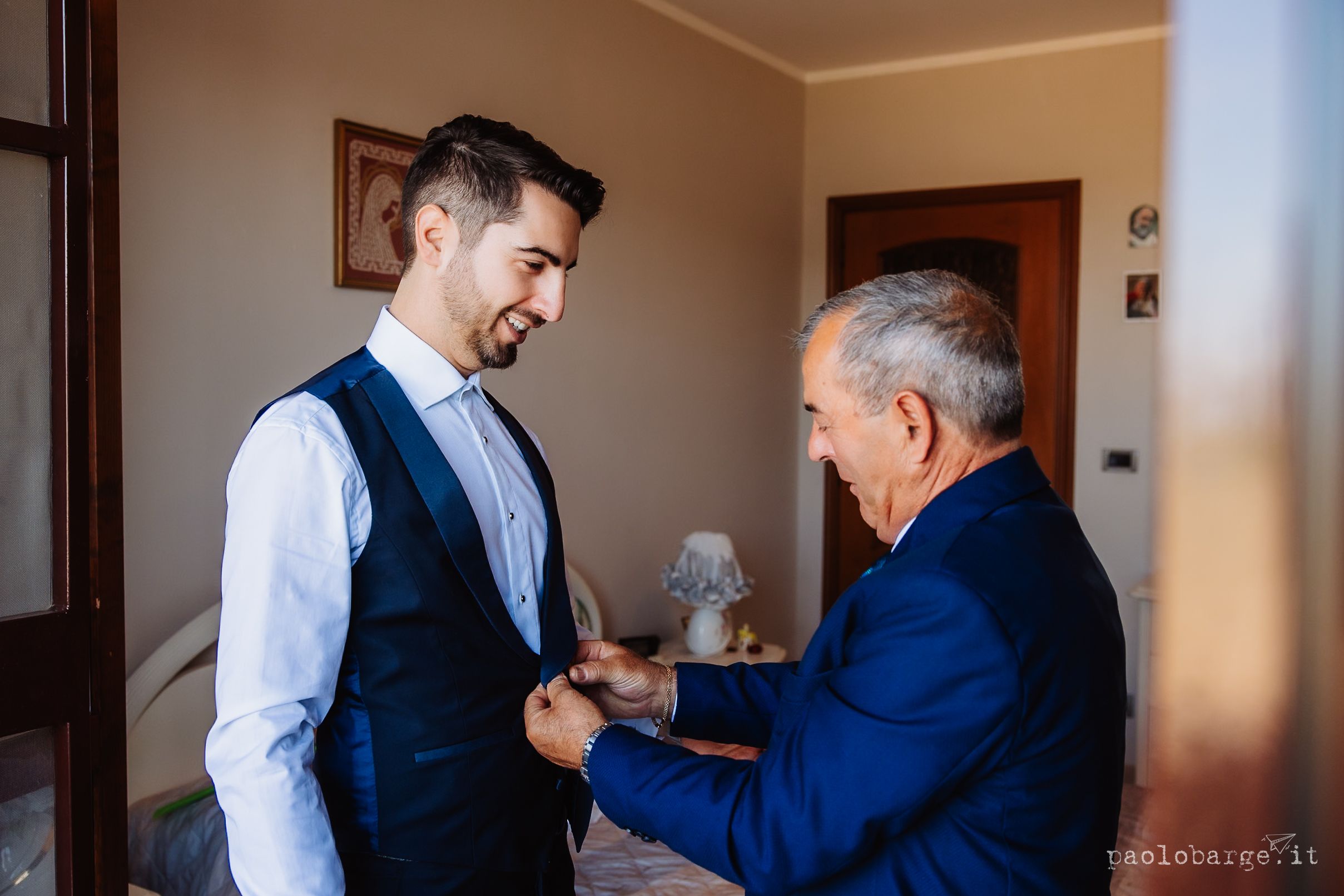 Two men stand together in a domestic setting while getting dressed in formal attire.