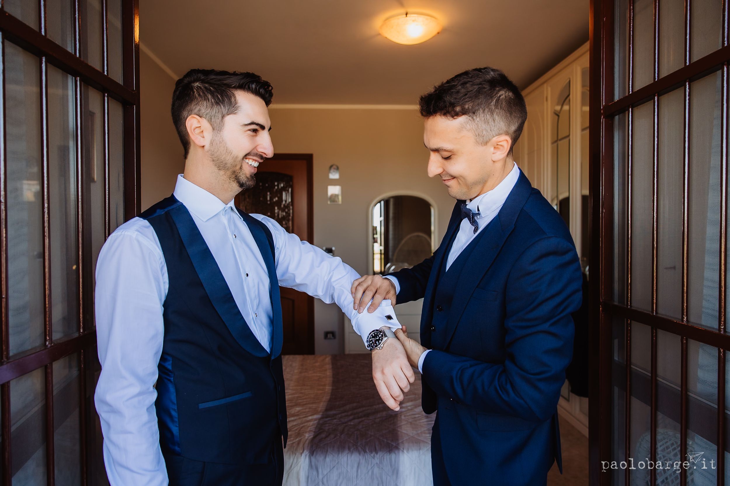 Two men in navy suits and light dress shirts share joyful moments in a hallway setting.