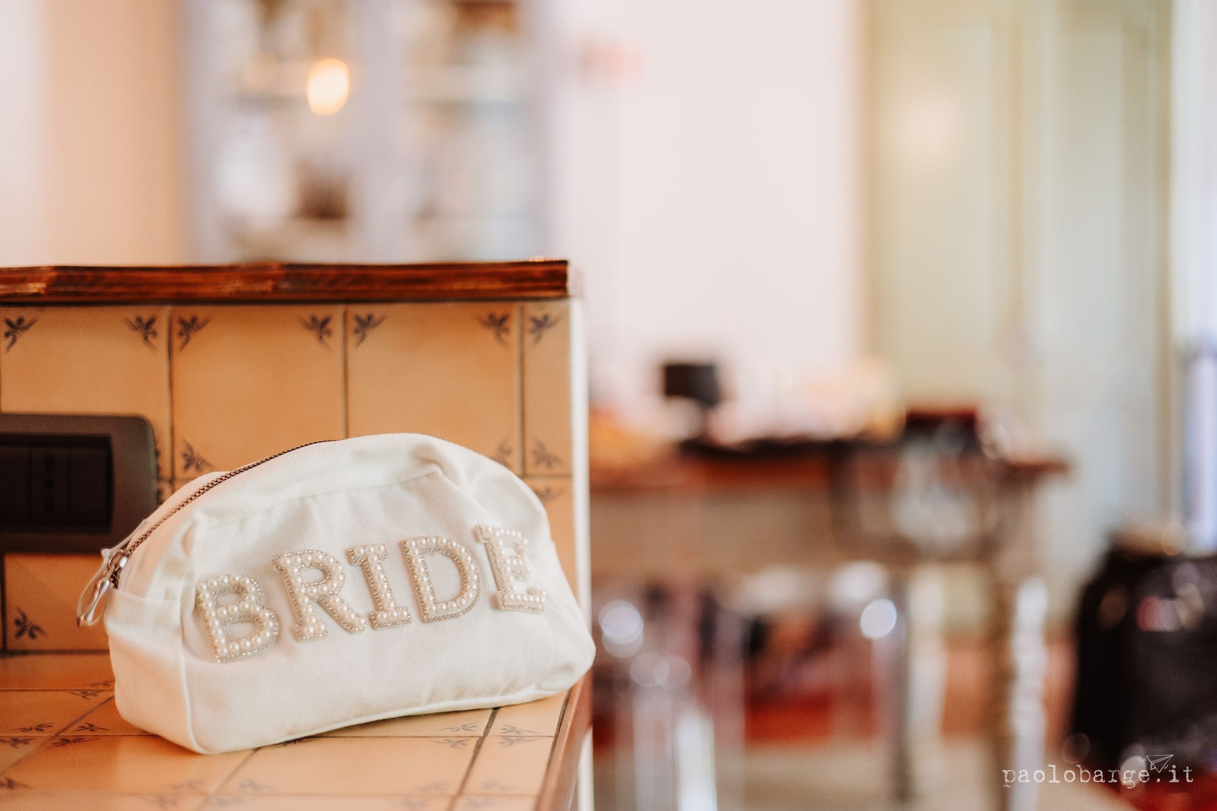 White cosmetic bag with 'BRIDE' text sits on wooden surface in soft focus interior setting.