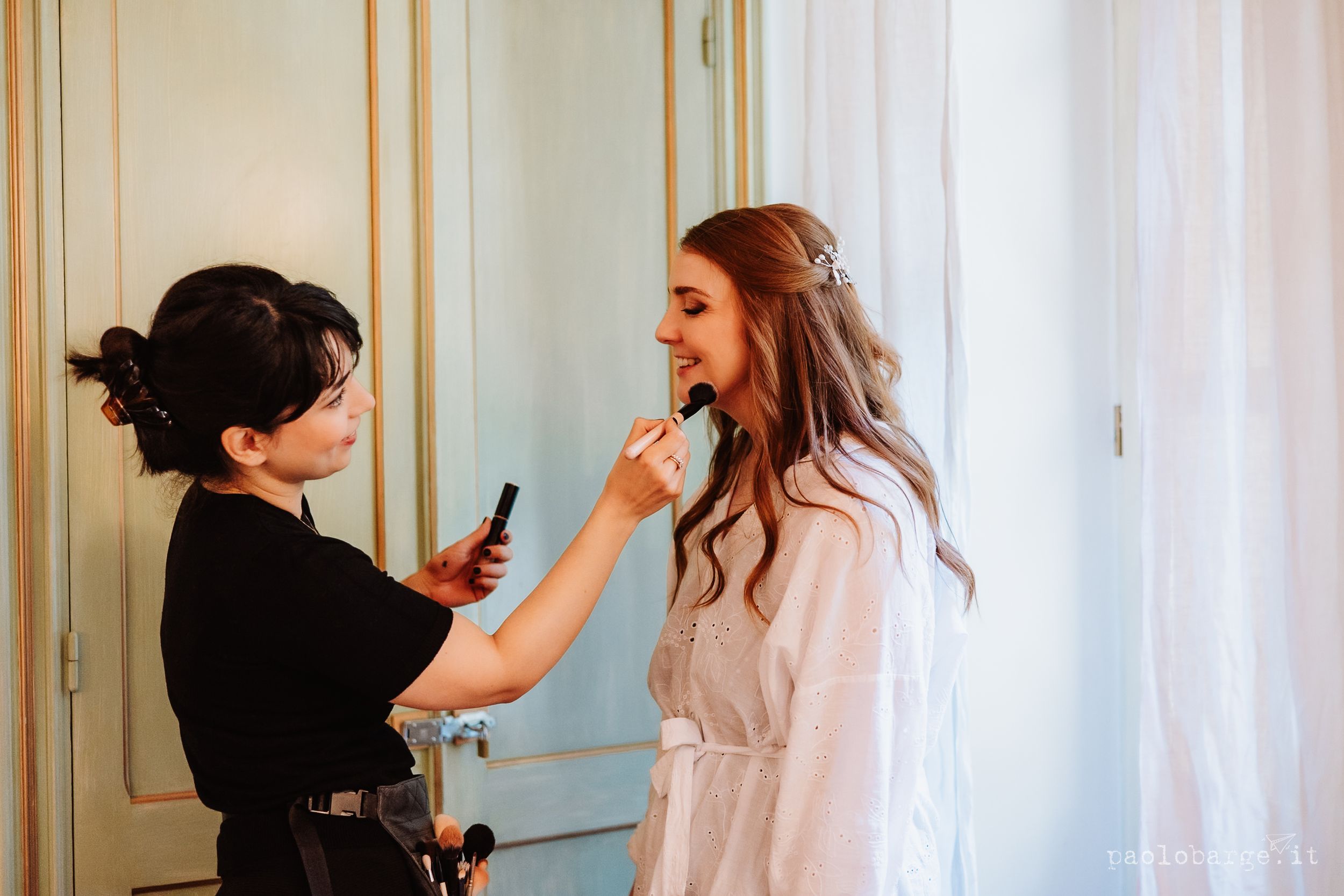 A makeup artist in black applies lipstick to a woman in a white robe with long wavy hair during a beauty session.