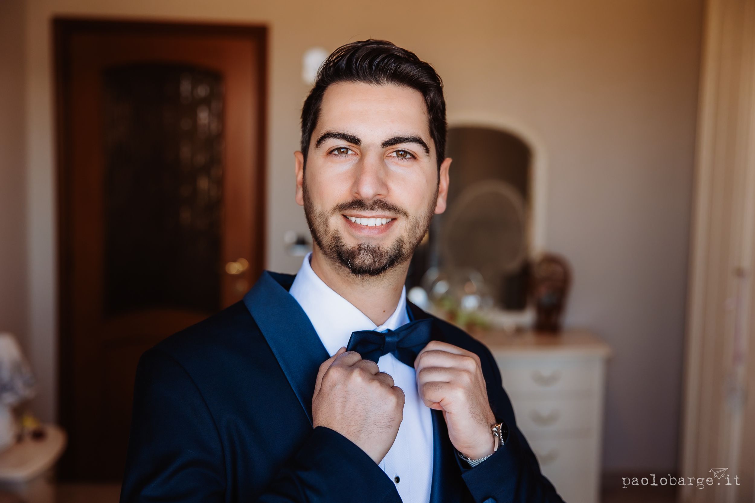 A smiling person in a navy blue suit adjusts their bowtie while getting ready indoors.
