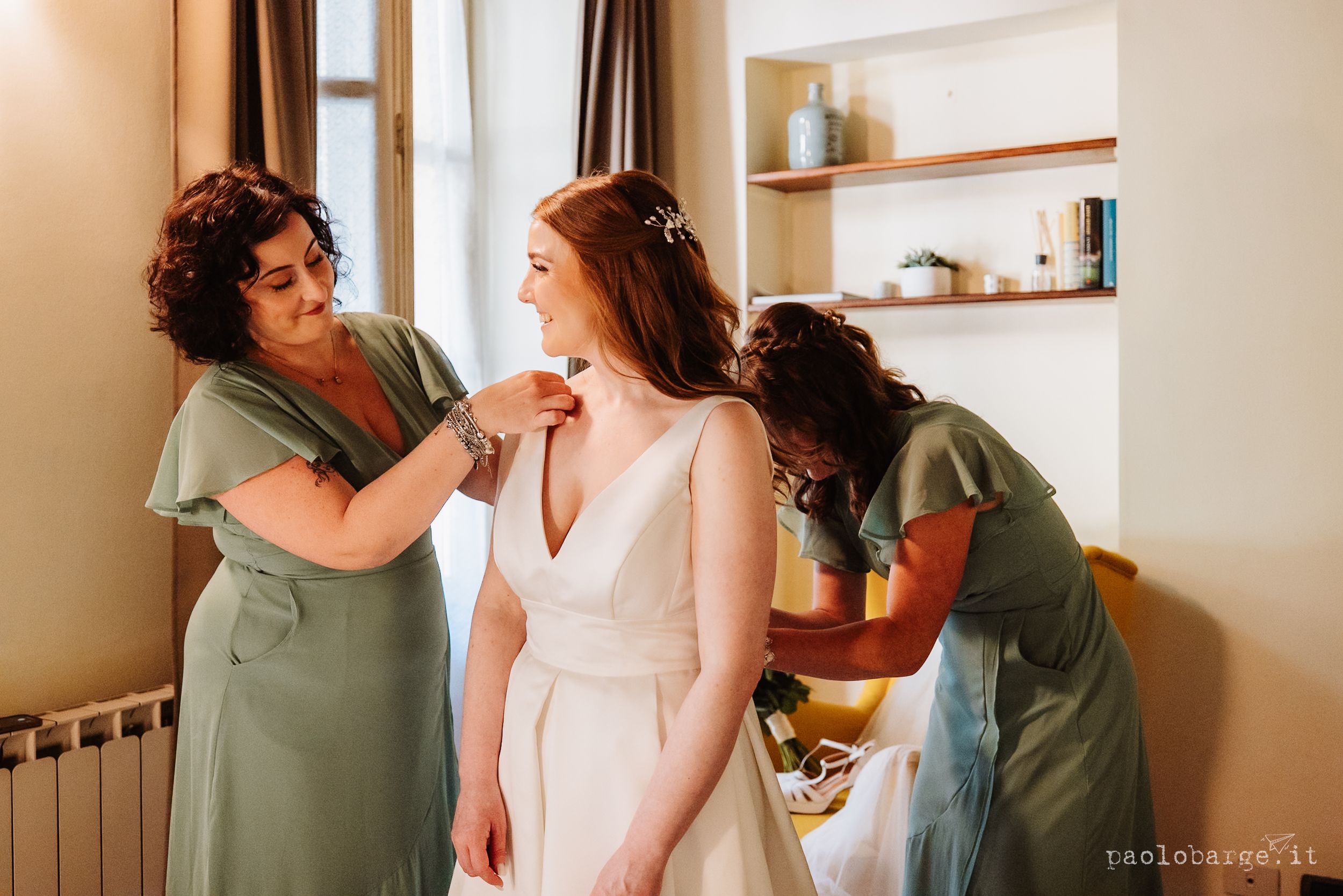 Two women share an intimate moment in a bright modern room with white walls and wooden shelf accents.