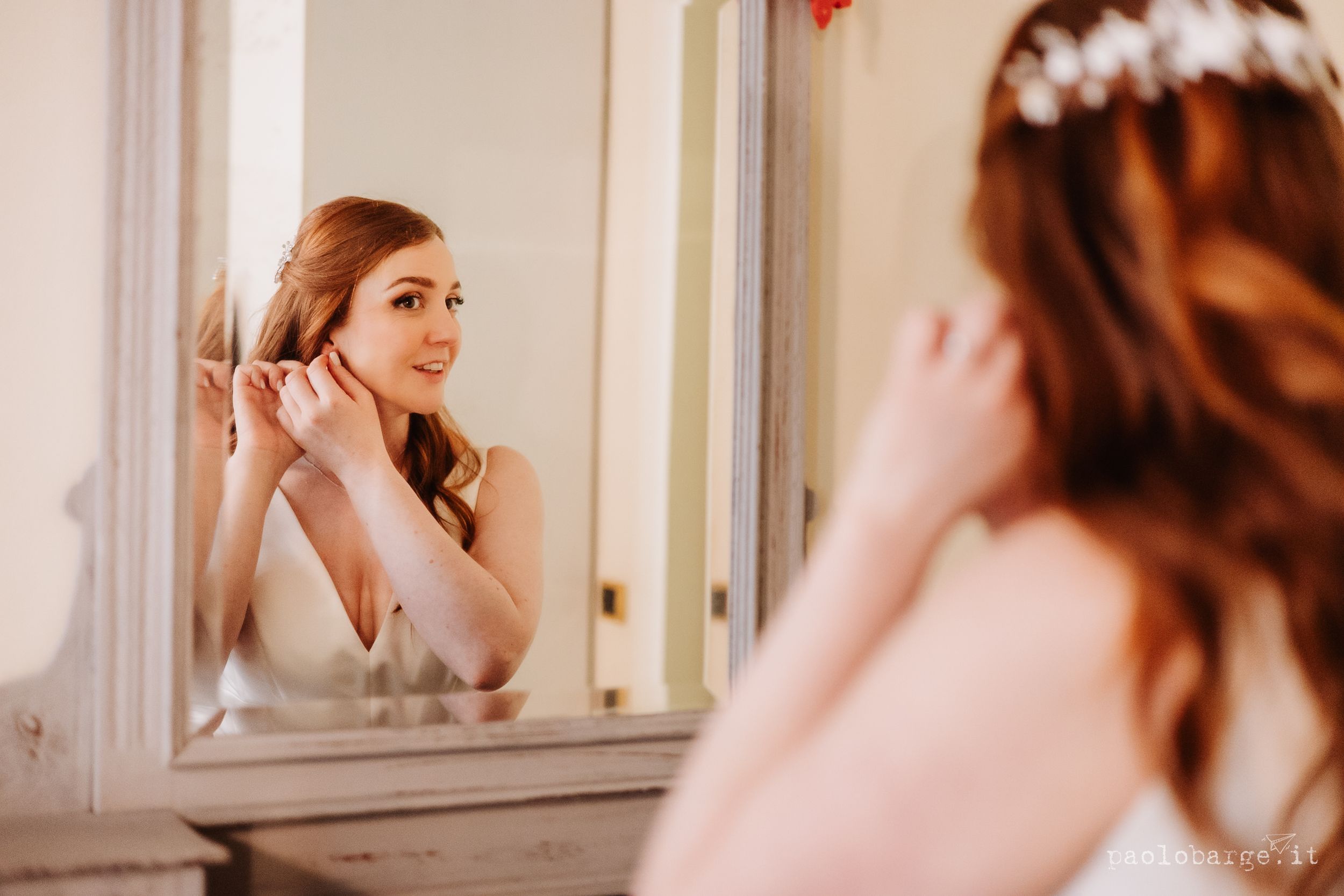 A bride admires her reflection in an elegant vintage mirror while putting on earrings during wedding preparations.