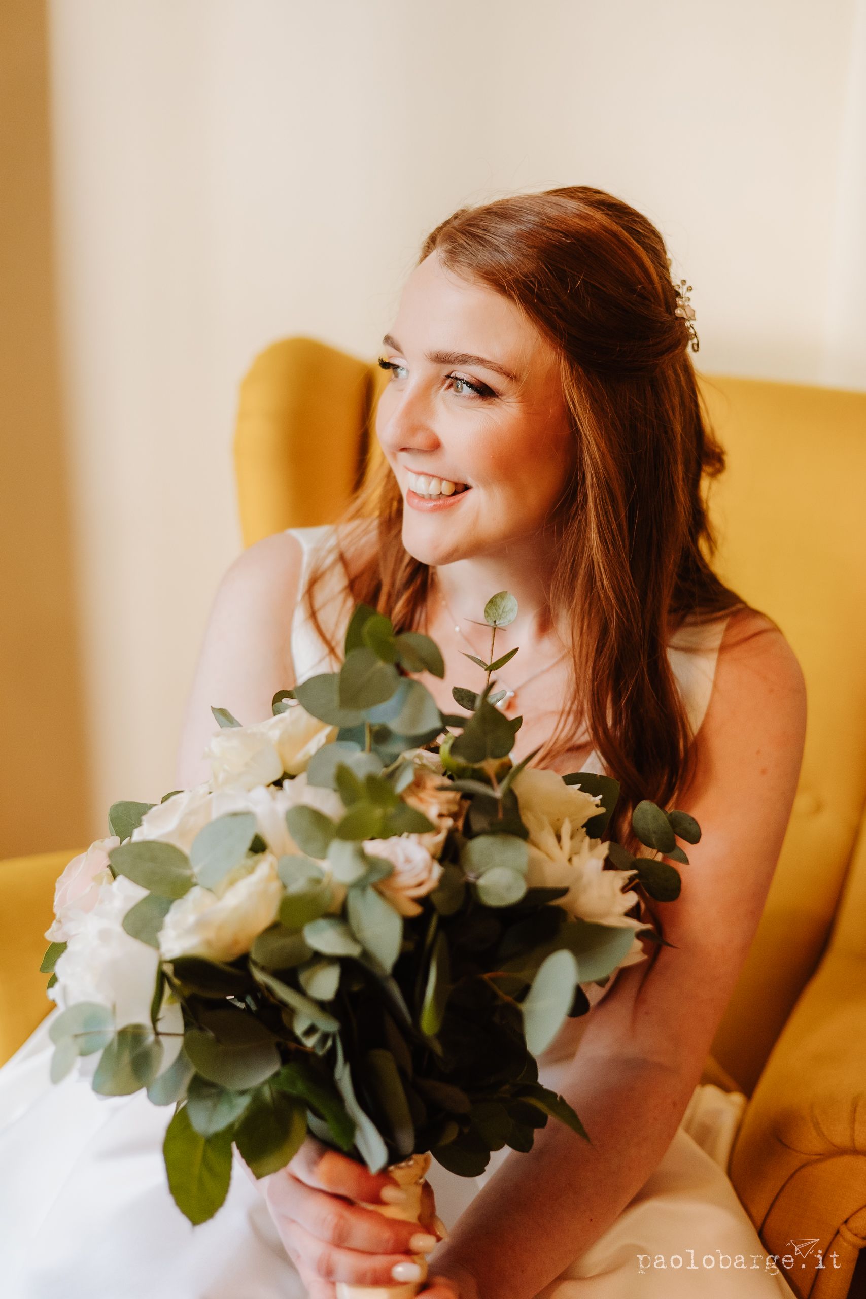 A bride in a white dress sits in a yellow chair holding a soft-toned bouquet with eucalyptus and white flowers.
