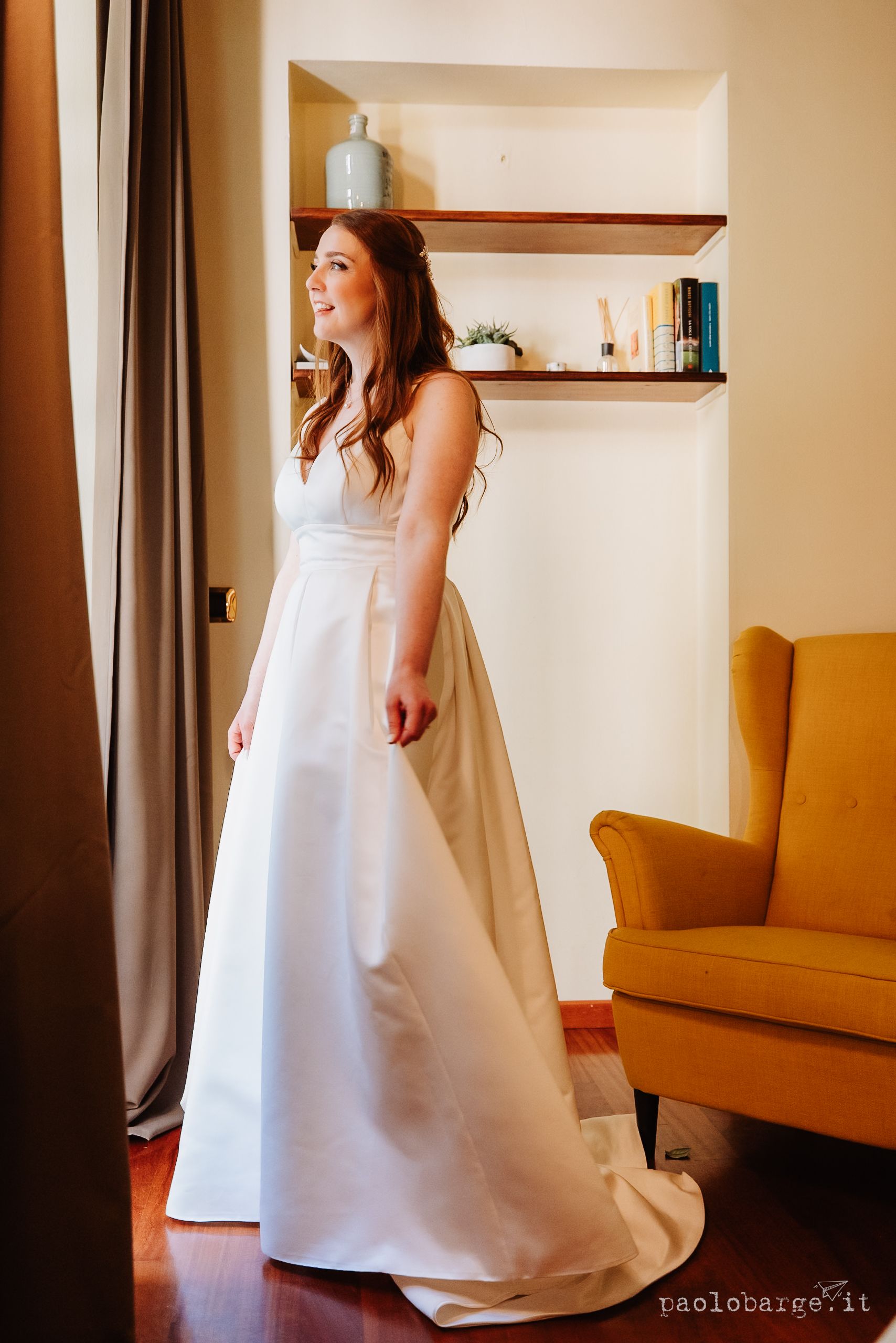 Inside a room with wooden shelves and a mustard yellow armchair, a person wears a flowing white wedding gown.