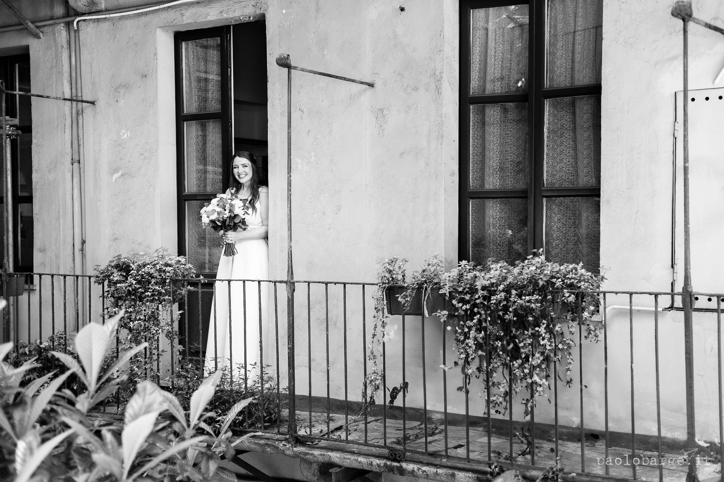 Black and white photo of a figure holding a bouquet on an elegant balcony with potted plants and railings.