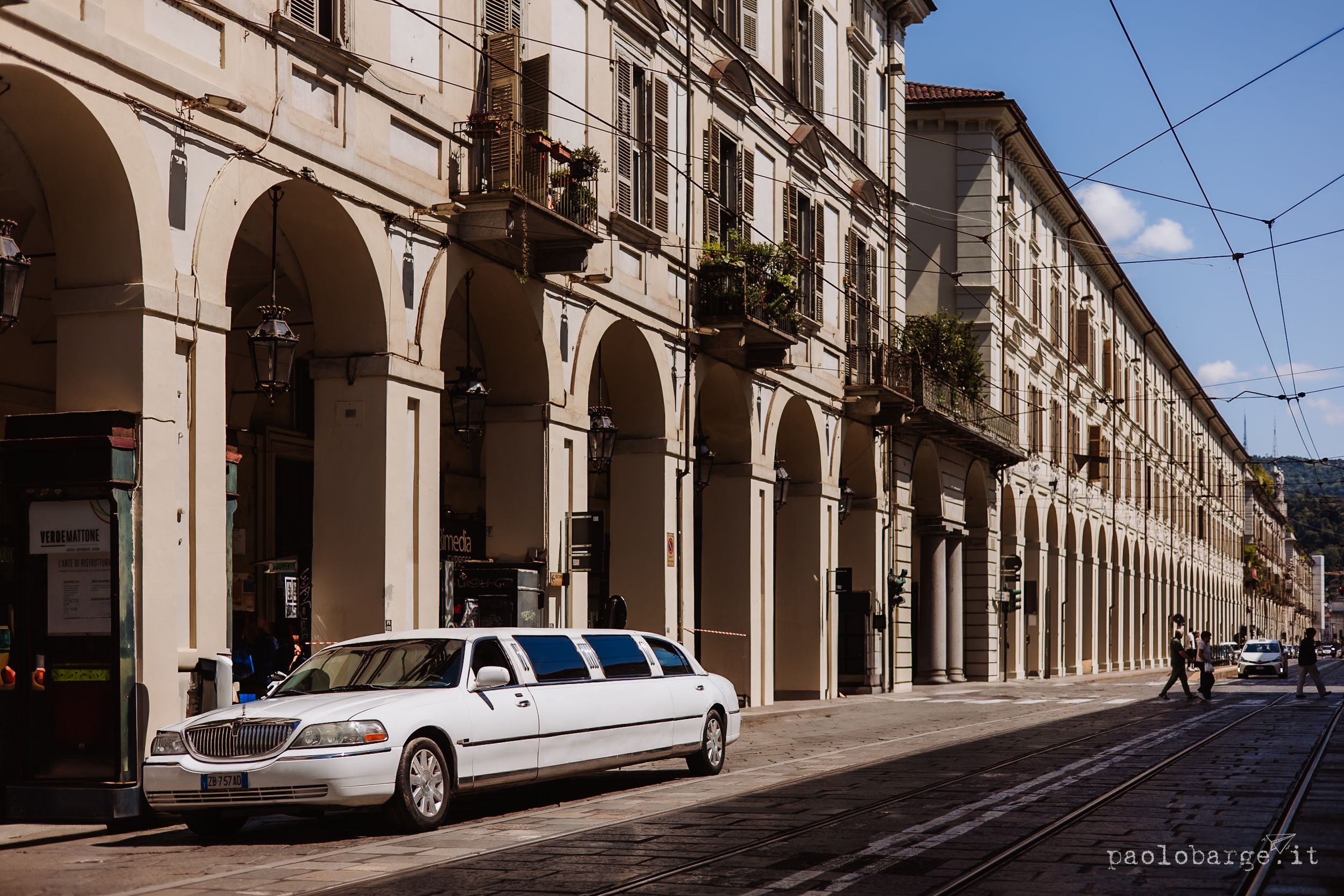 A white stretch limousine parked along a historic arcade street with elegant stone archways in Turin, Italy.