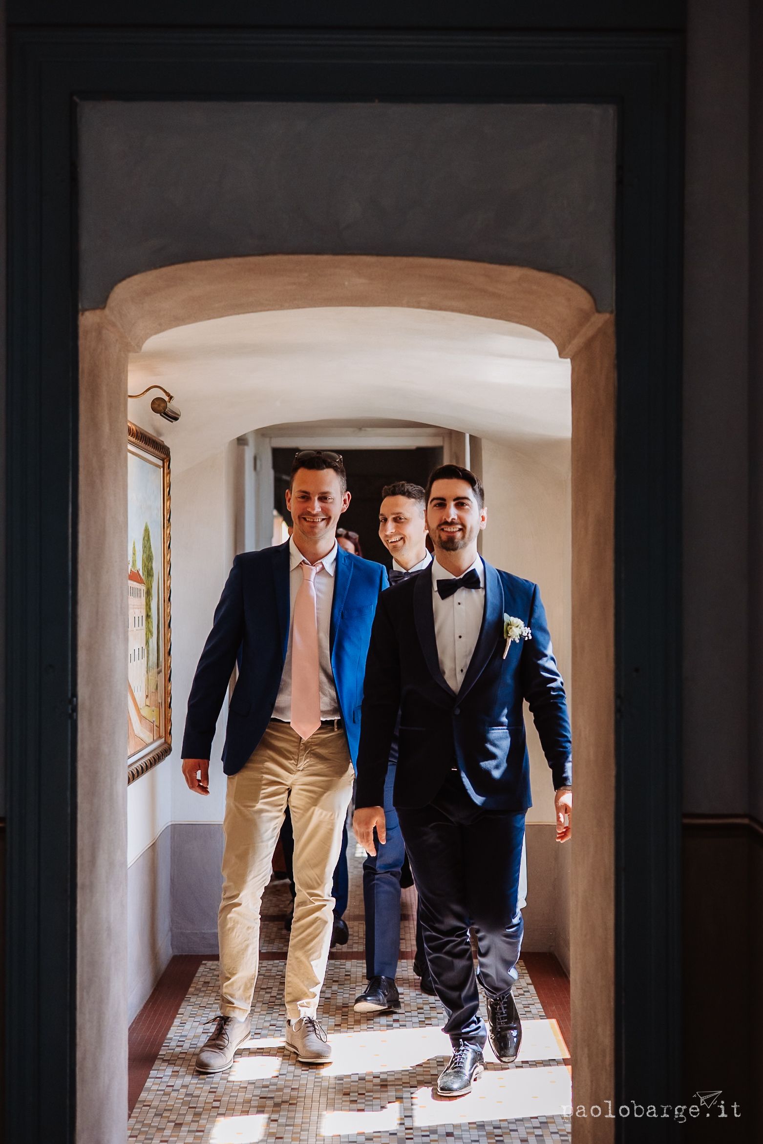 Three men in formal attire walk through an arched hallway during a wedding celebration.