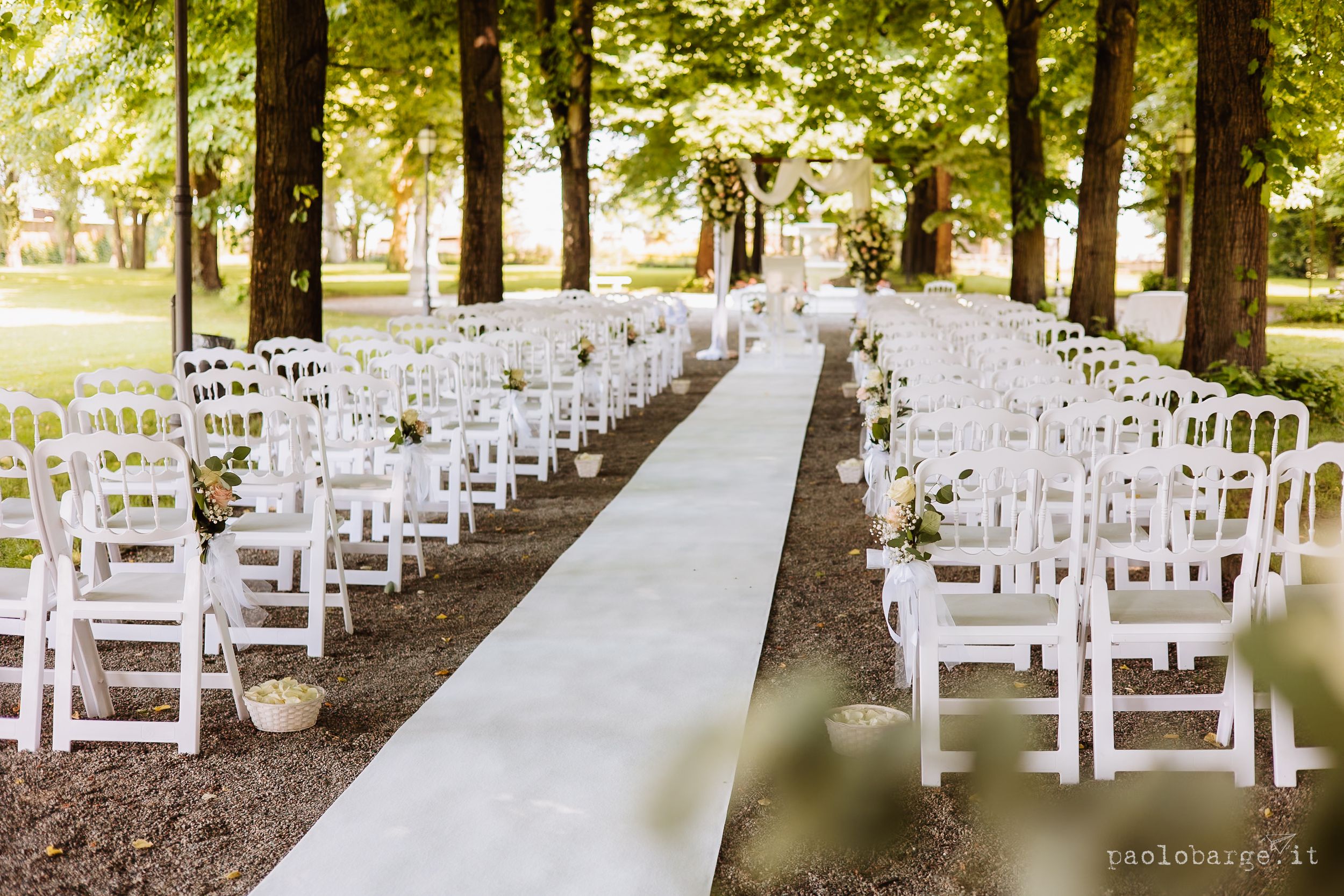 White chairs line both sides of an aisle runner beneath tall trees for an outdoor wedding ceremony setup.