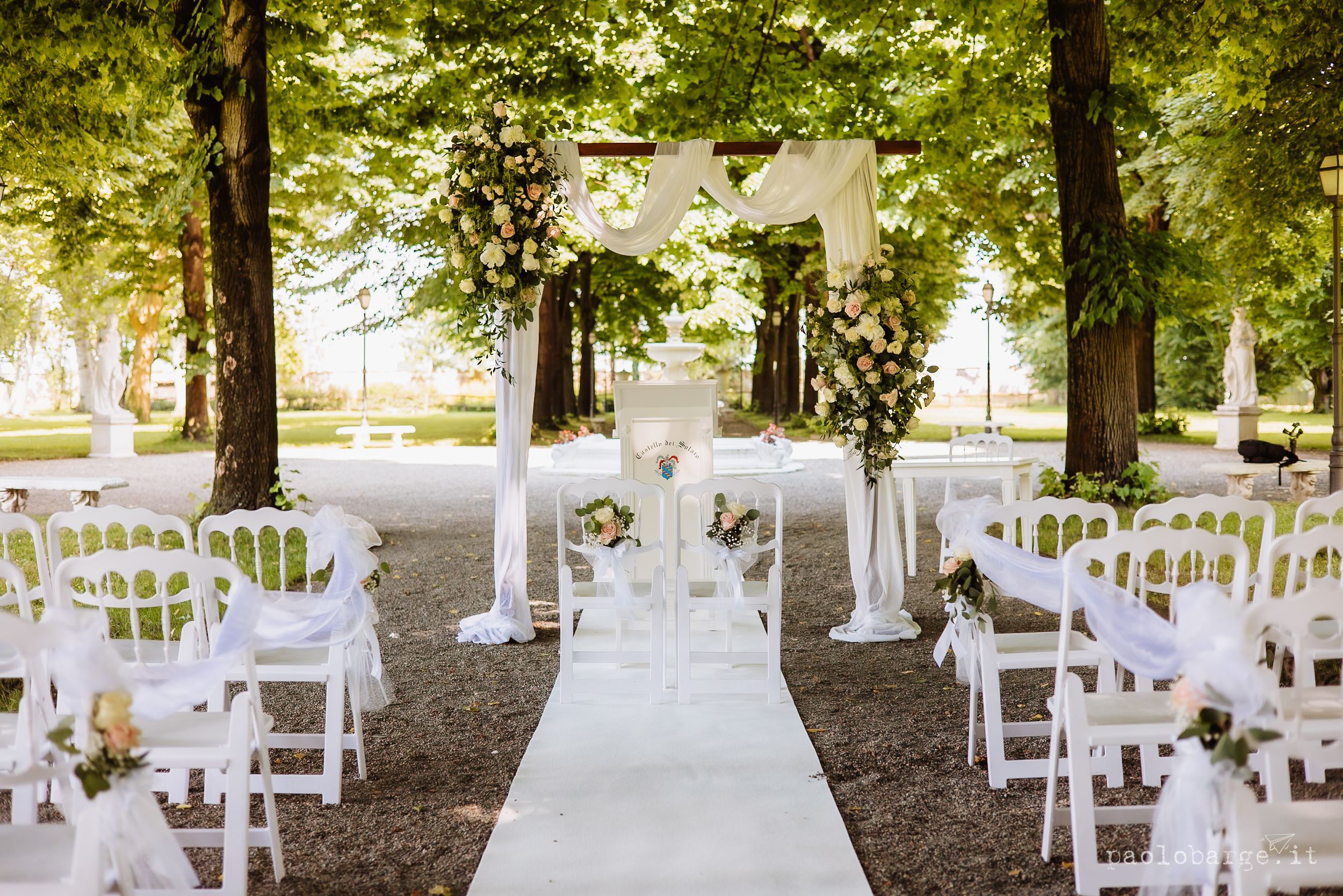 White chairs line a floral-decorated aisle path in an outdoor garden wedding ceremony setup under shaded trees.
