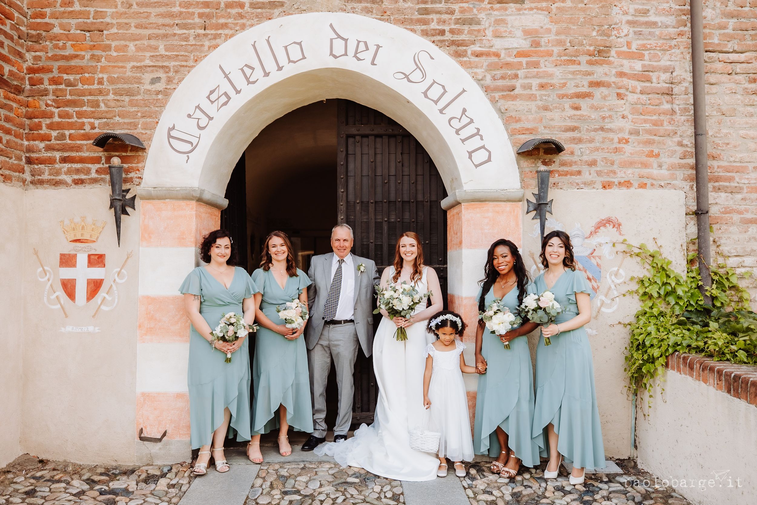 A wedding party in light blue dresses poses beneath a stone archway entrance at Castello del Solaro.