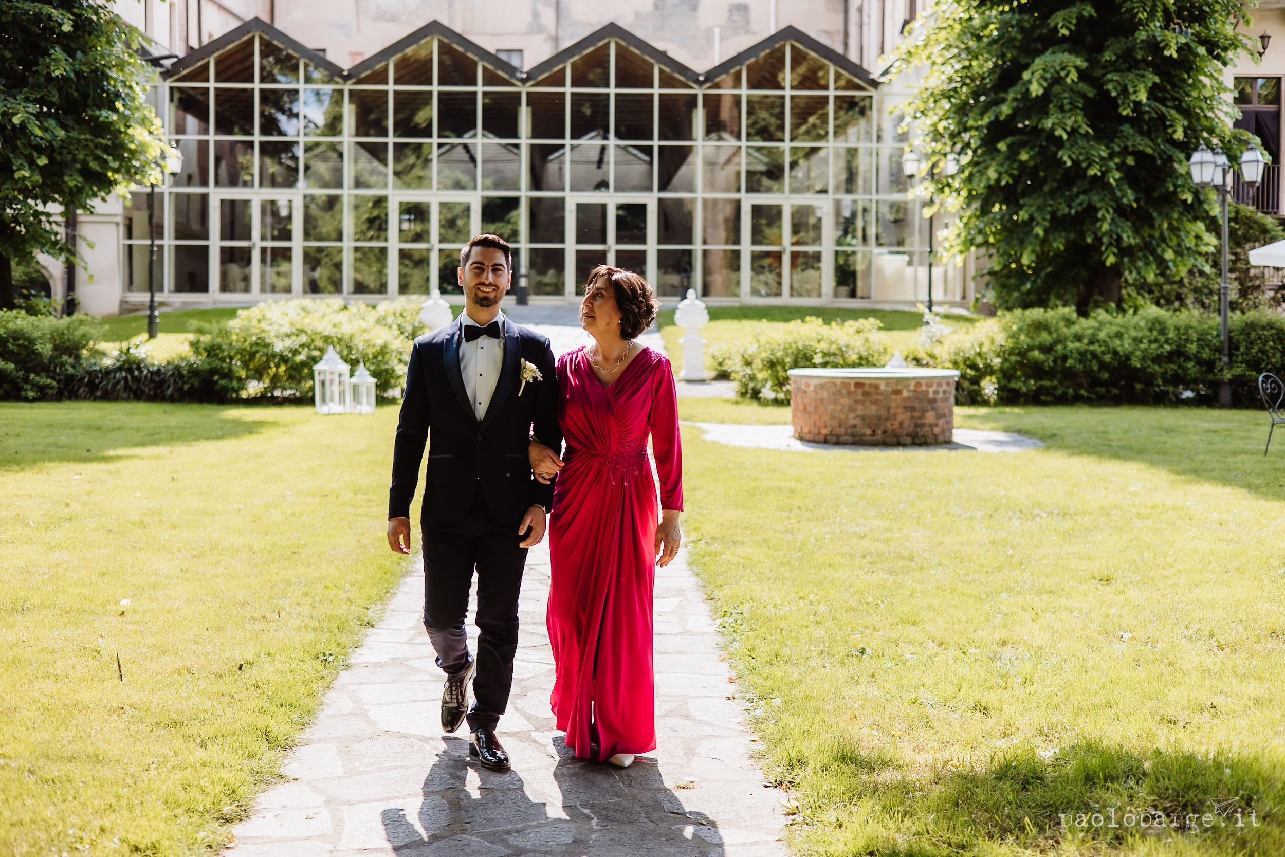 A couple walks along a stone pathway through lush green gardens at an elegant wedding venue.