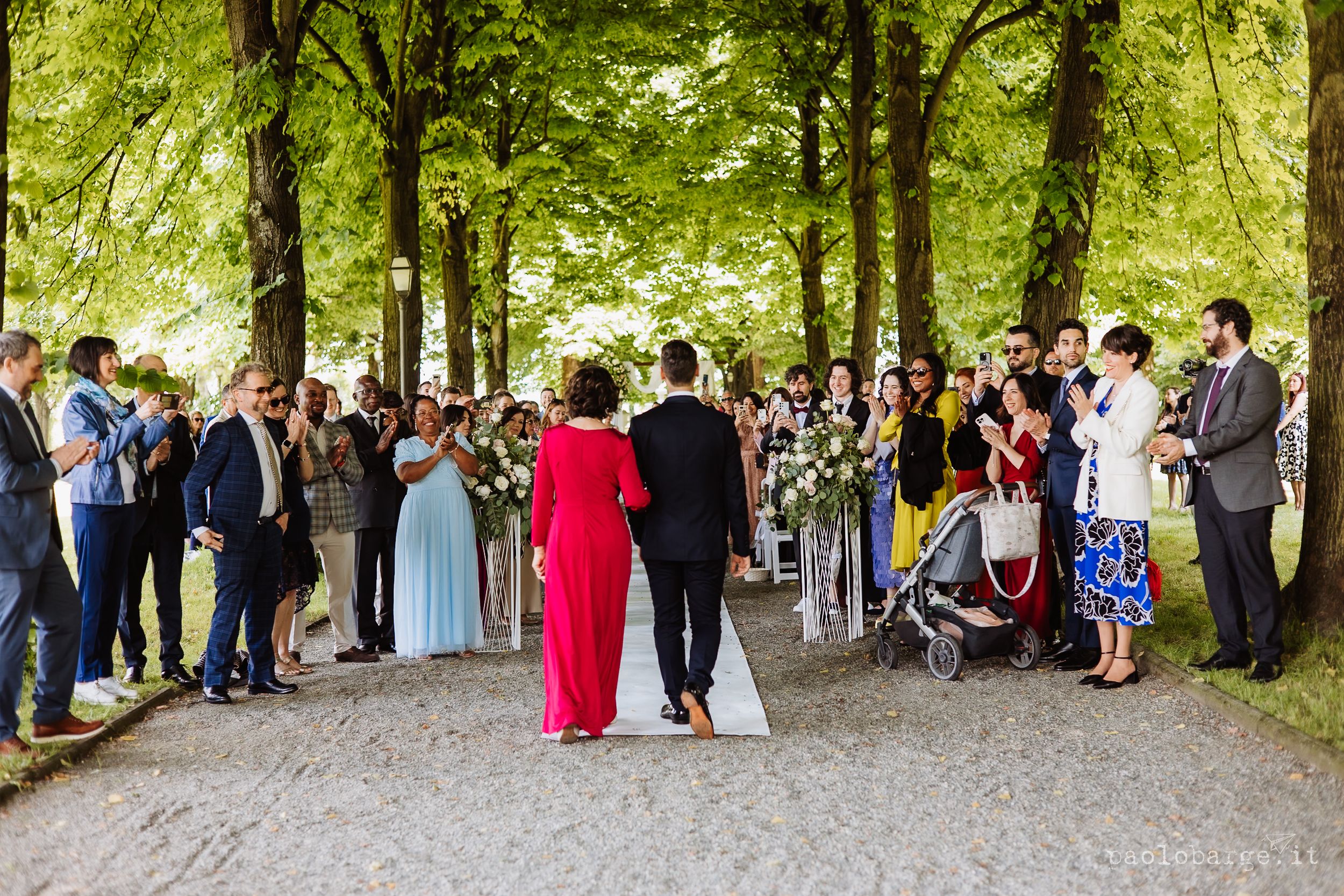 Wedding guests gather beneath green trees in a park setting for an outdoor celebration.