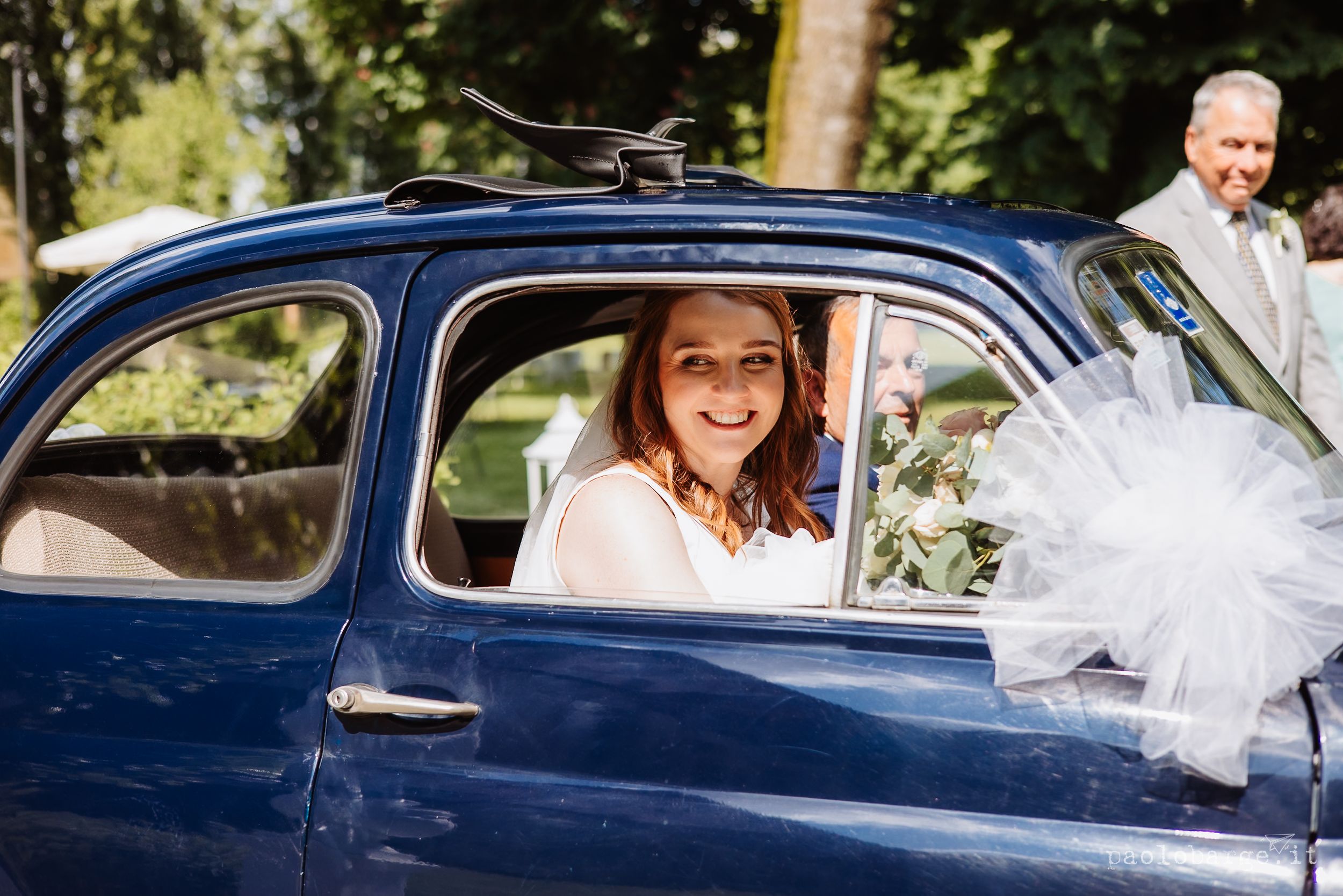 A classic blue vintage car decorated with flowers drives down a sunny road with happy passengers inside.