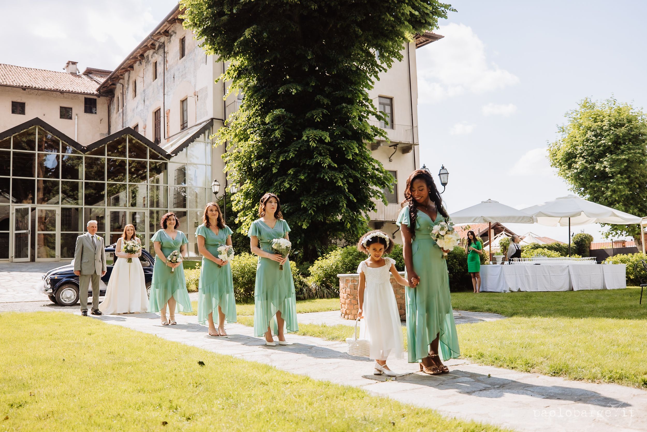 Wedding party wearing mint green dresses poses joyfully on a grassy hillside on a sunny summer day.