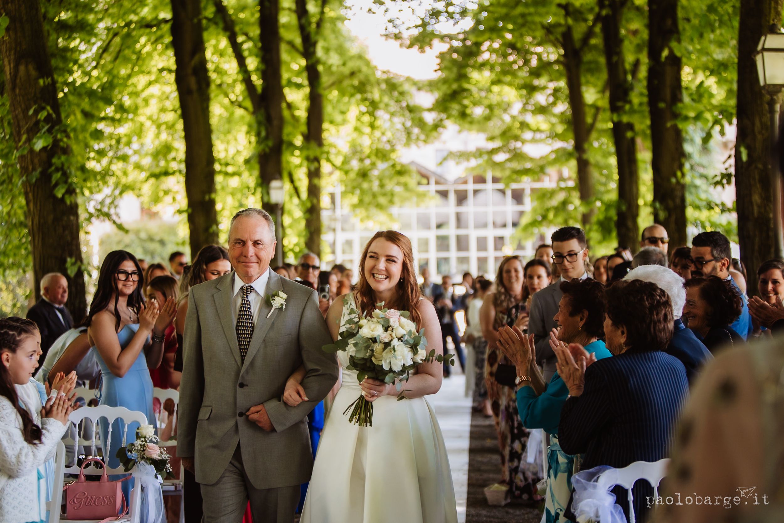 Outdoor wedding ceremony with tall trees and guests gathered as couple exchanges vows.