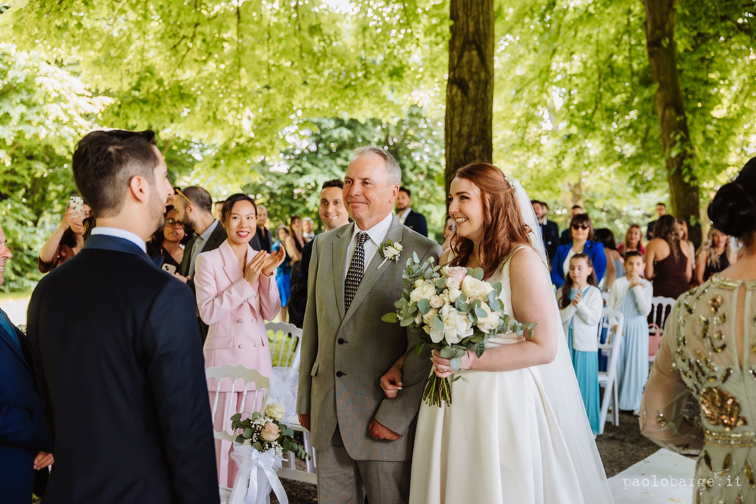 Wedding guests gather outdoors at a sunny garden reception with elegant white floral arrangements and natural decor.