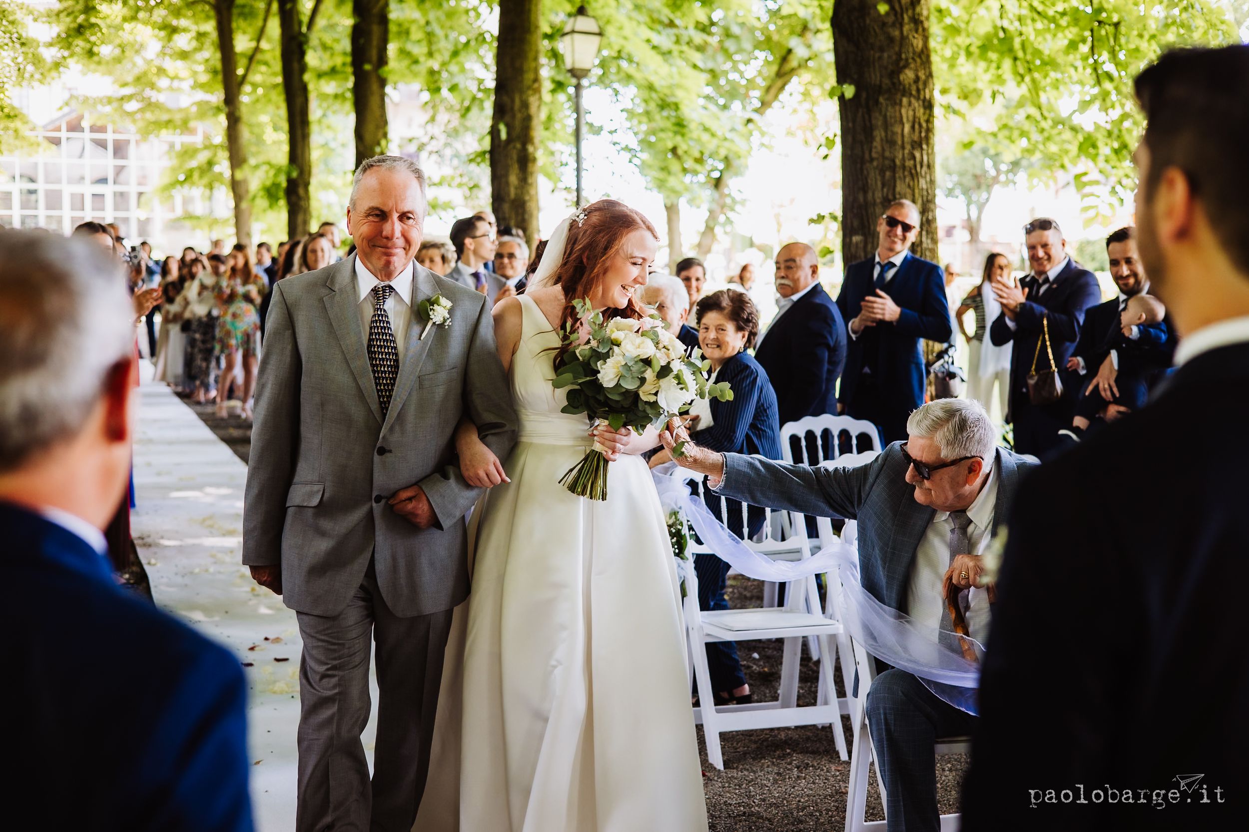 Elegant outdoor wedding ceremony features guests seated in white chairs under a canopy of green trees in a garden setting.