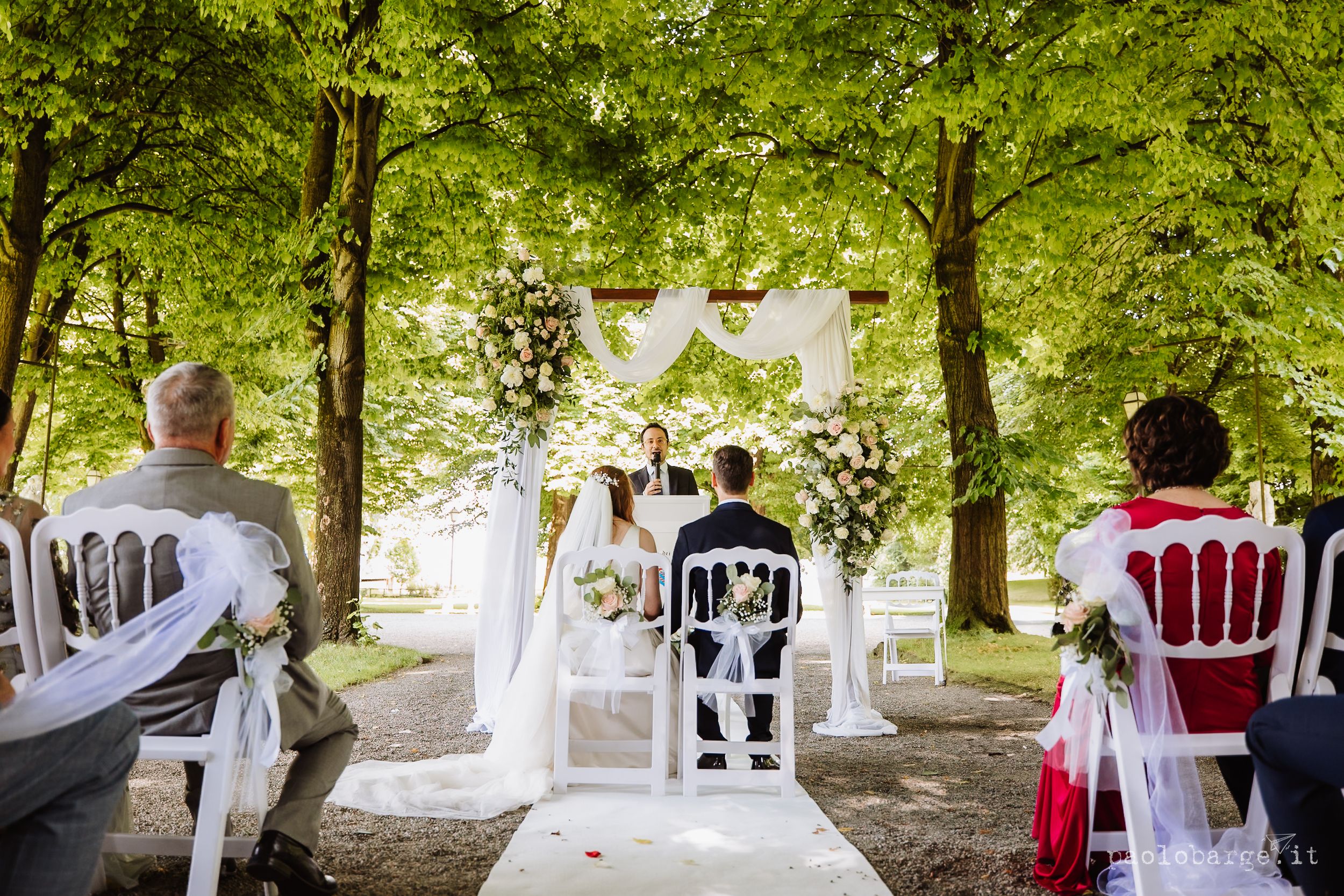 White outdoor wedding reception setup under trees with plastic chairs and fluttering fabric decorations.