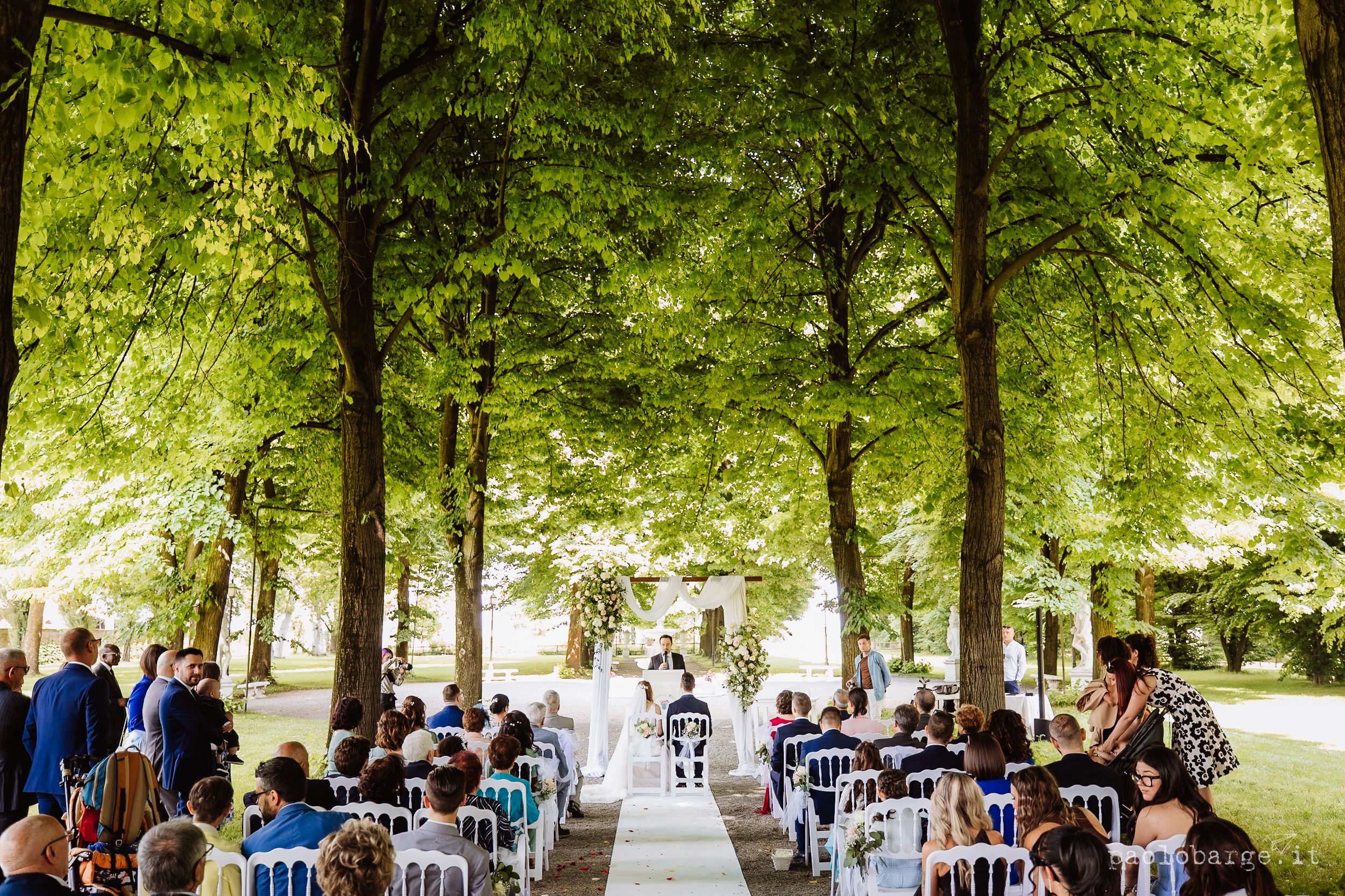 A rustic outdoor wedding ceremony set under tall shady trees with rows of white chairs and natural decor.