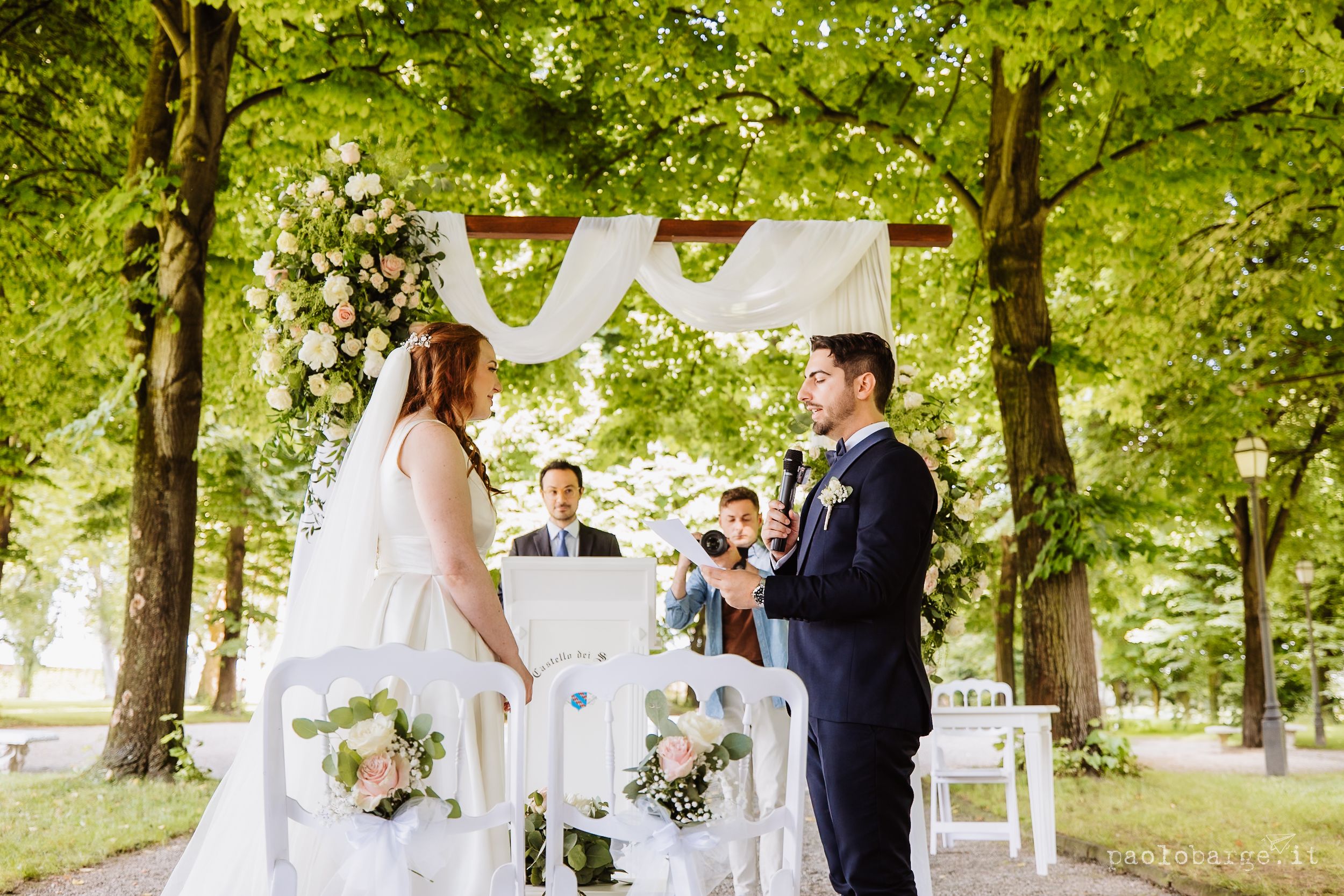 Outdoor wedding ceremony under leafy green trees with white decor and floral arrangements.
