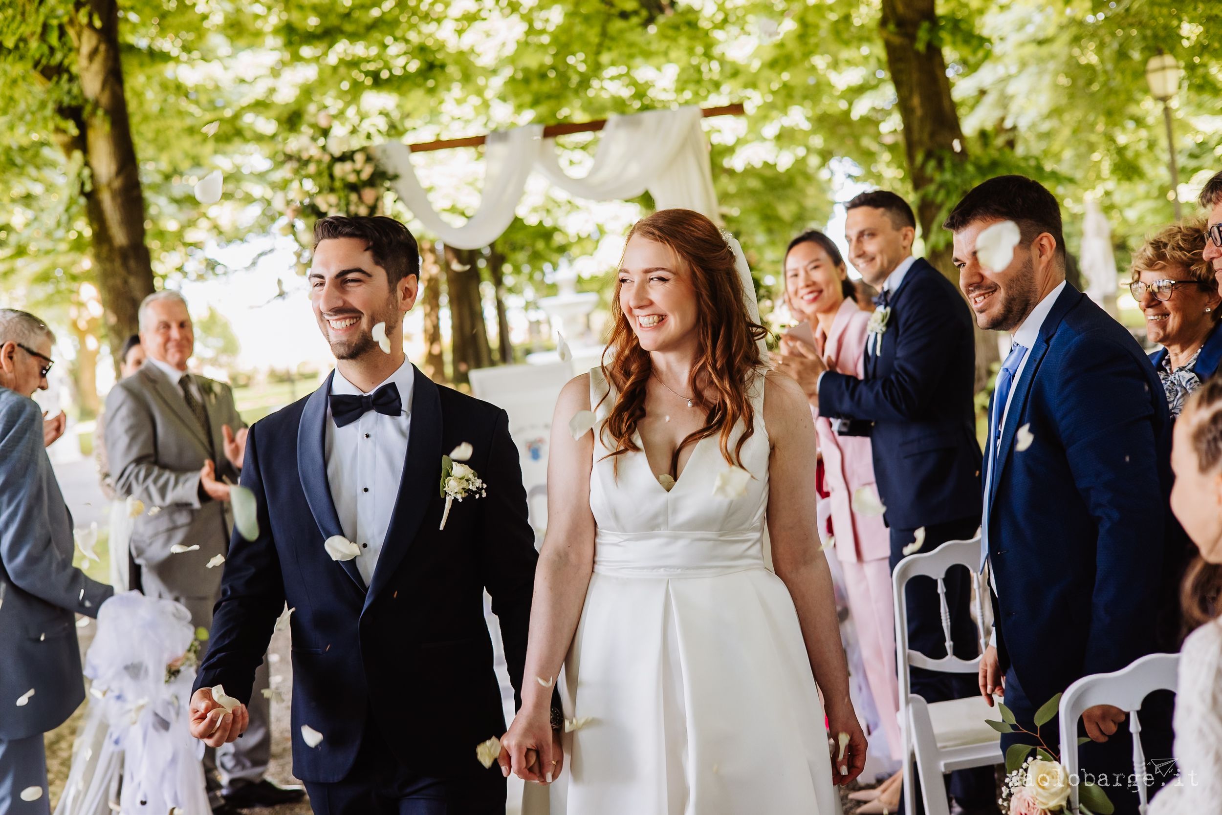 Happy wedding guests mingle and socialize at an outdoor reception under a canopy of green trees.