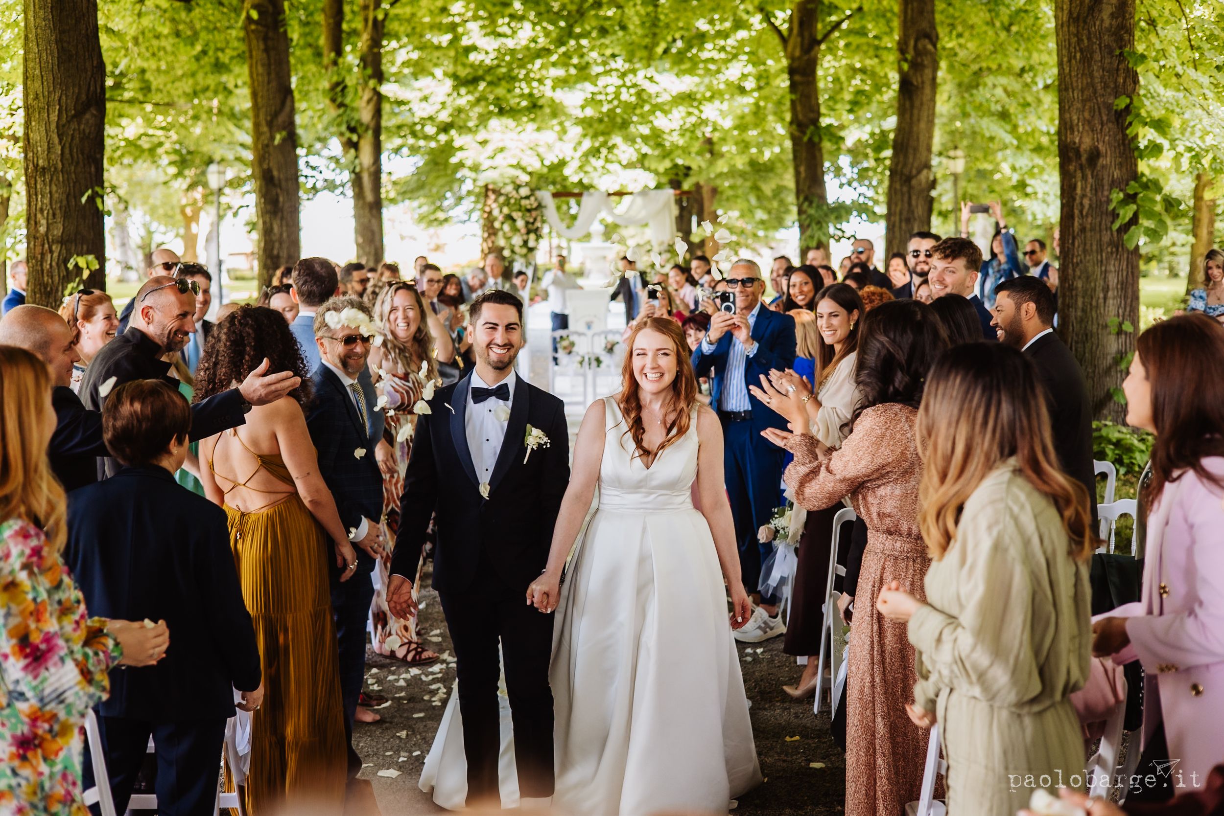 Wedding couple walks down tree-lined path surrounded by celebrating guests on their special day outdoors.