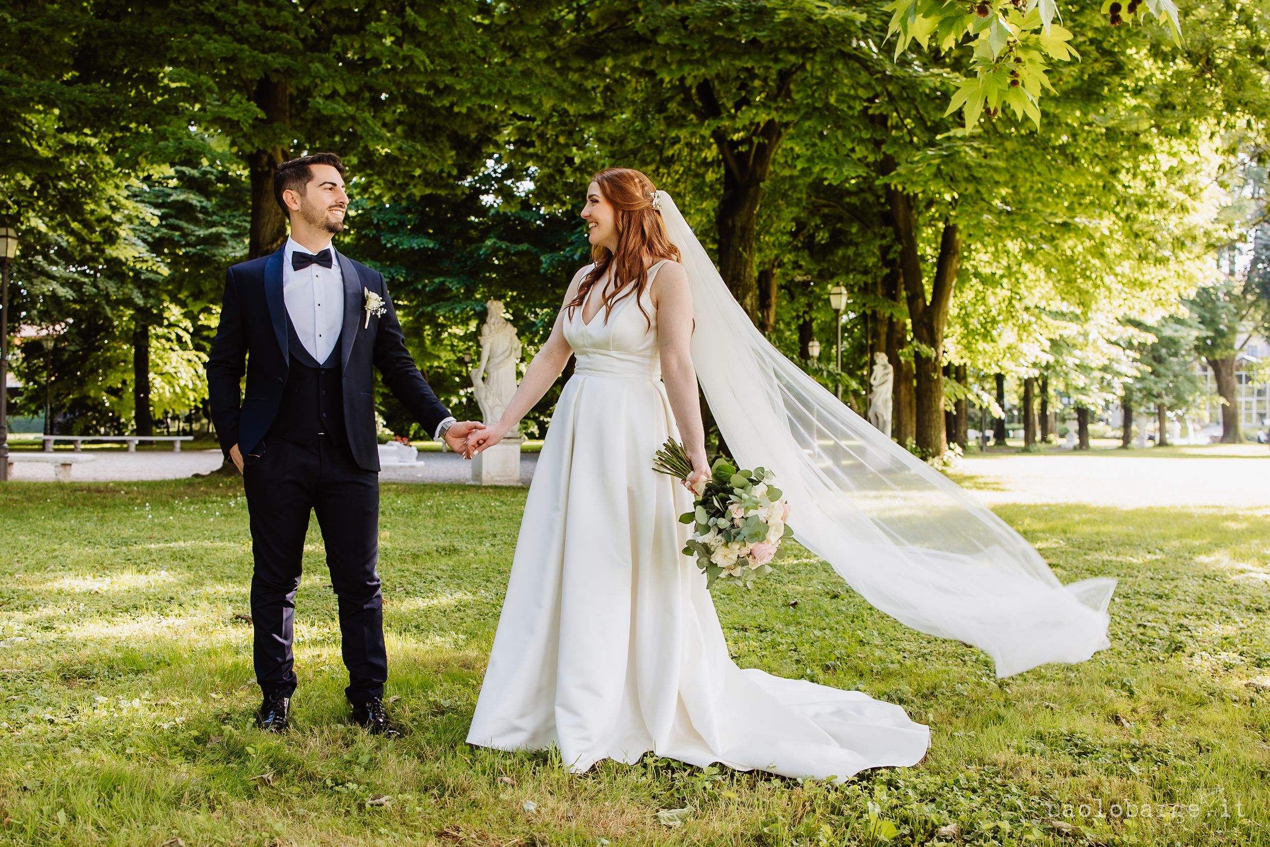 Wedding couple walks together through sunlit park surrounded by lush green trees on sunny summer afternoon.