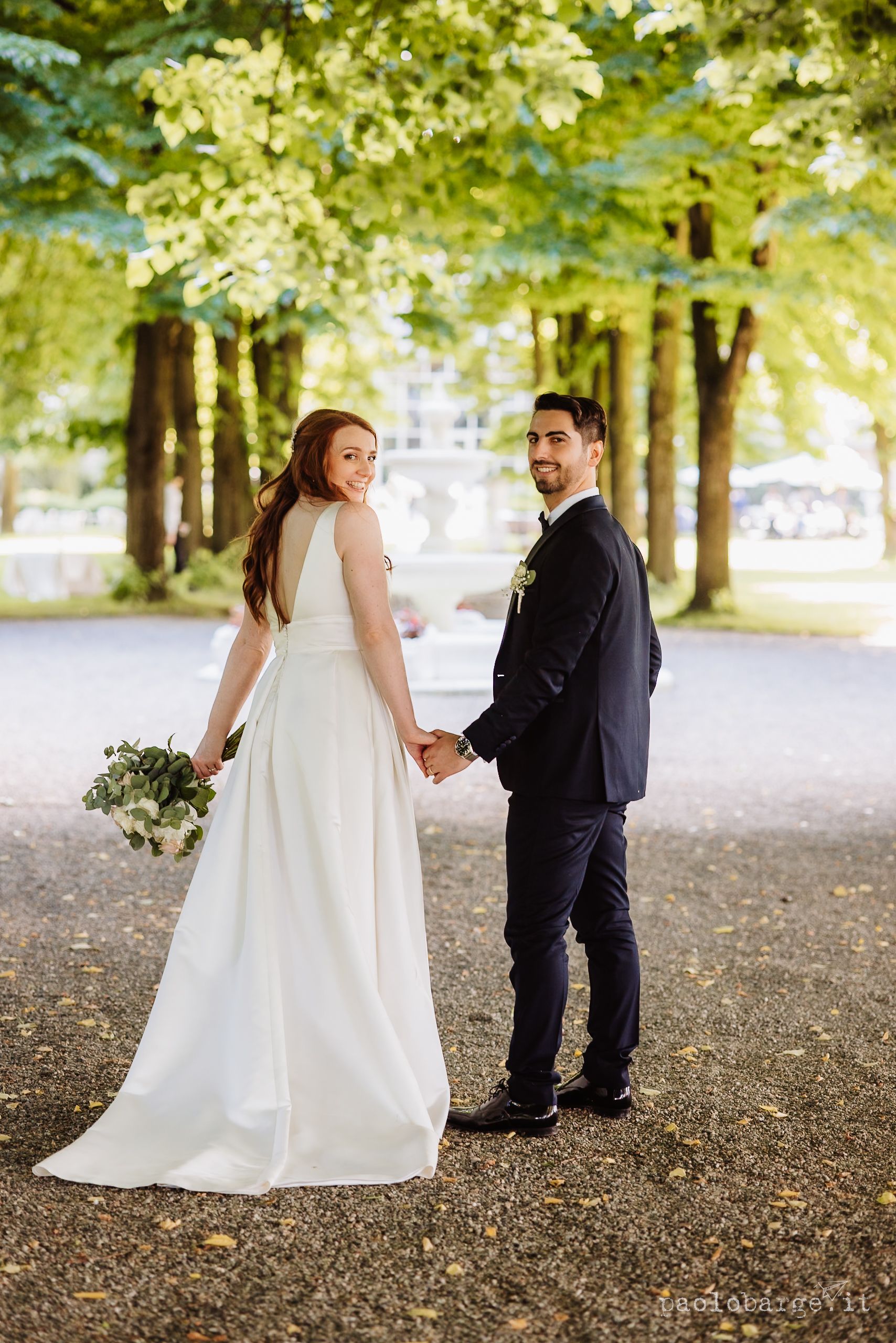 A couple in wedding attire holding hands and looking back at the camera while walking under tree-lined path in park.