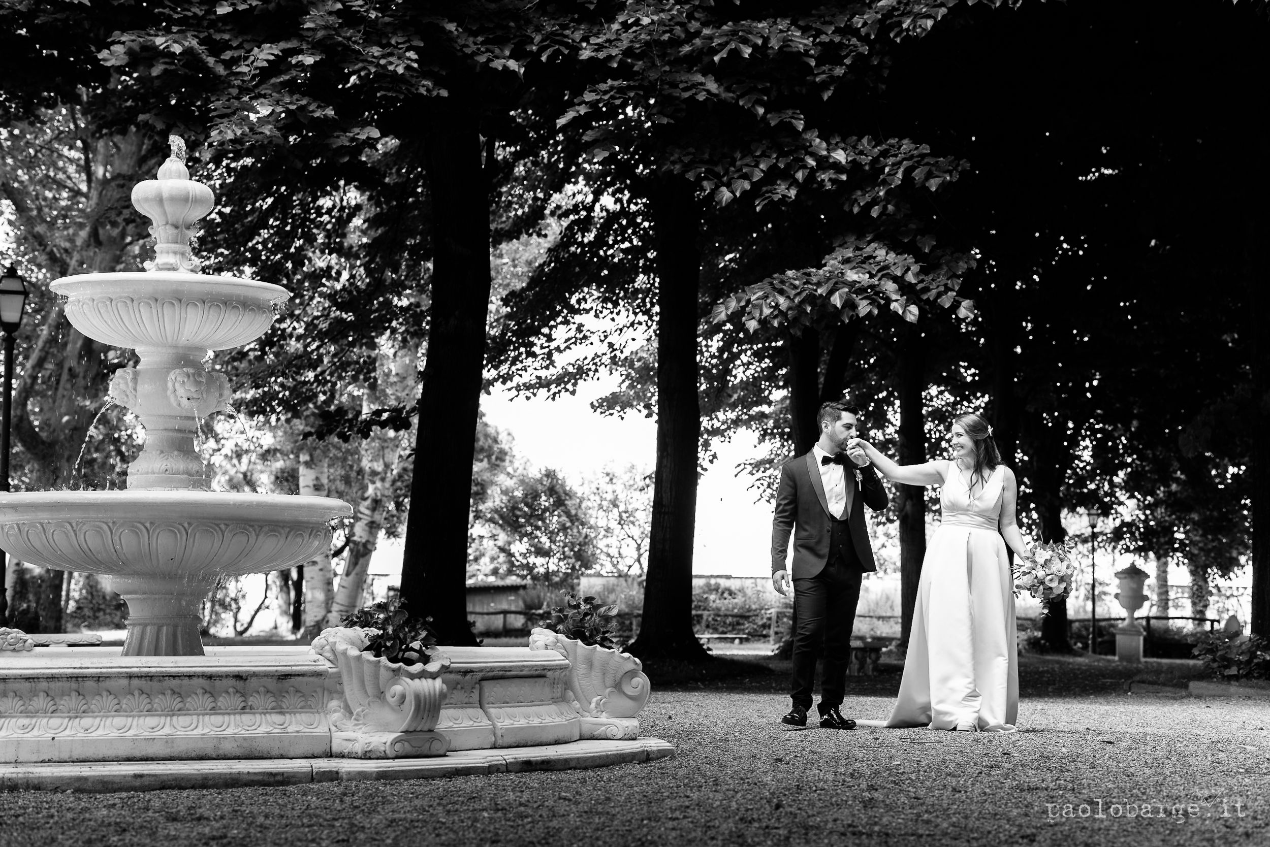 A black and white vintage photograph captures an elegant couple standing near an ornate garden fountain in a park.