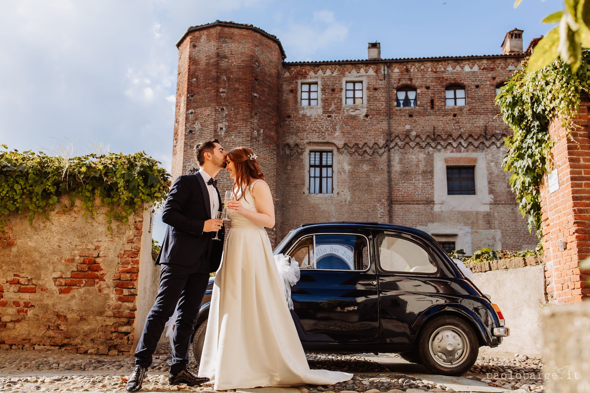 A romantic outdoor wedding scene with a classic vintage car and rustic brick architecture in warm sunlight.