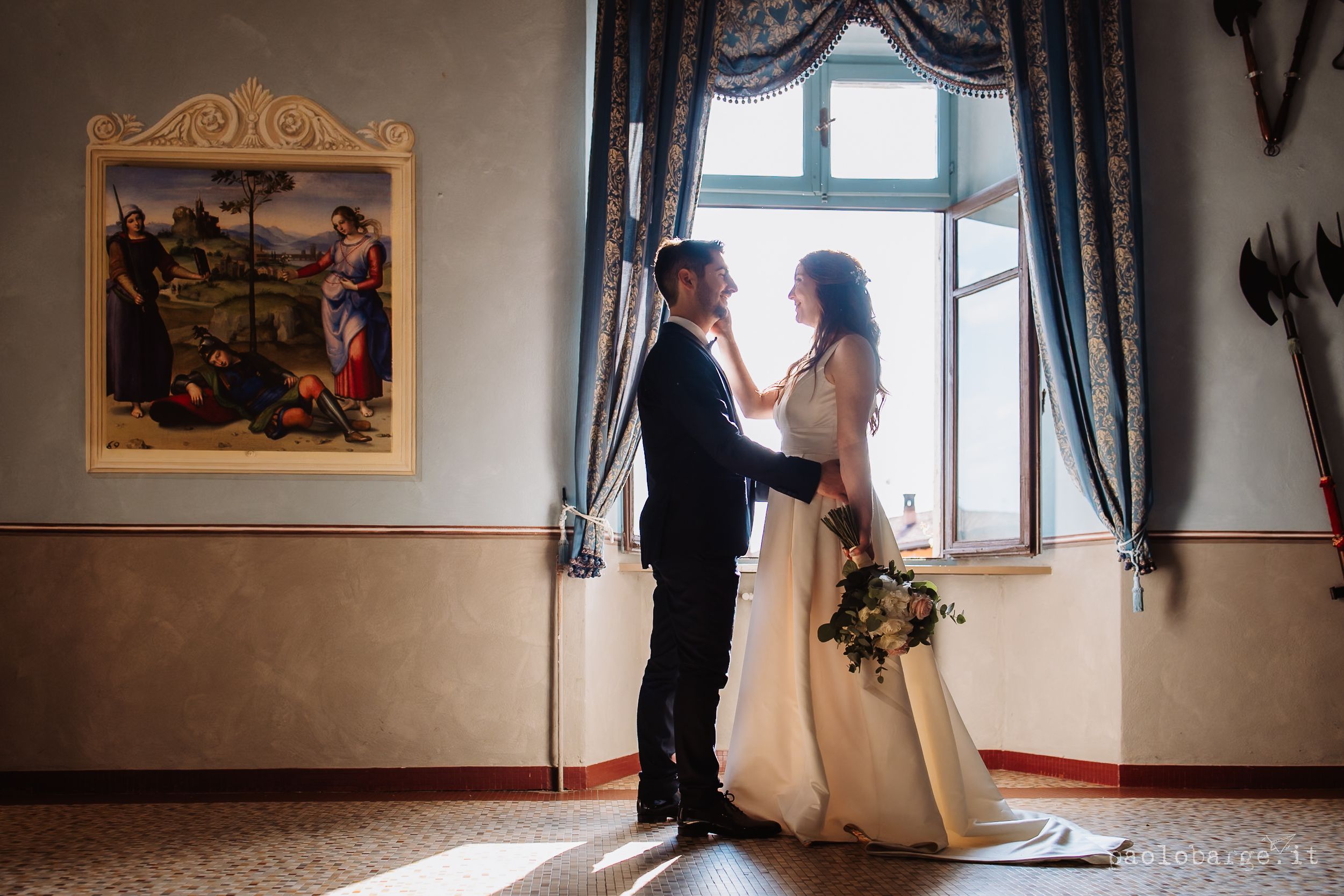 A couple stands near a decorative window in a historic building interior as light streams through the glass.