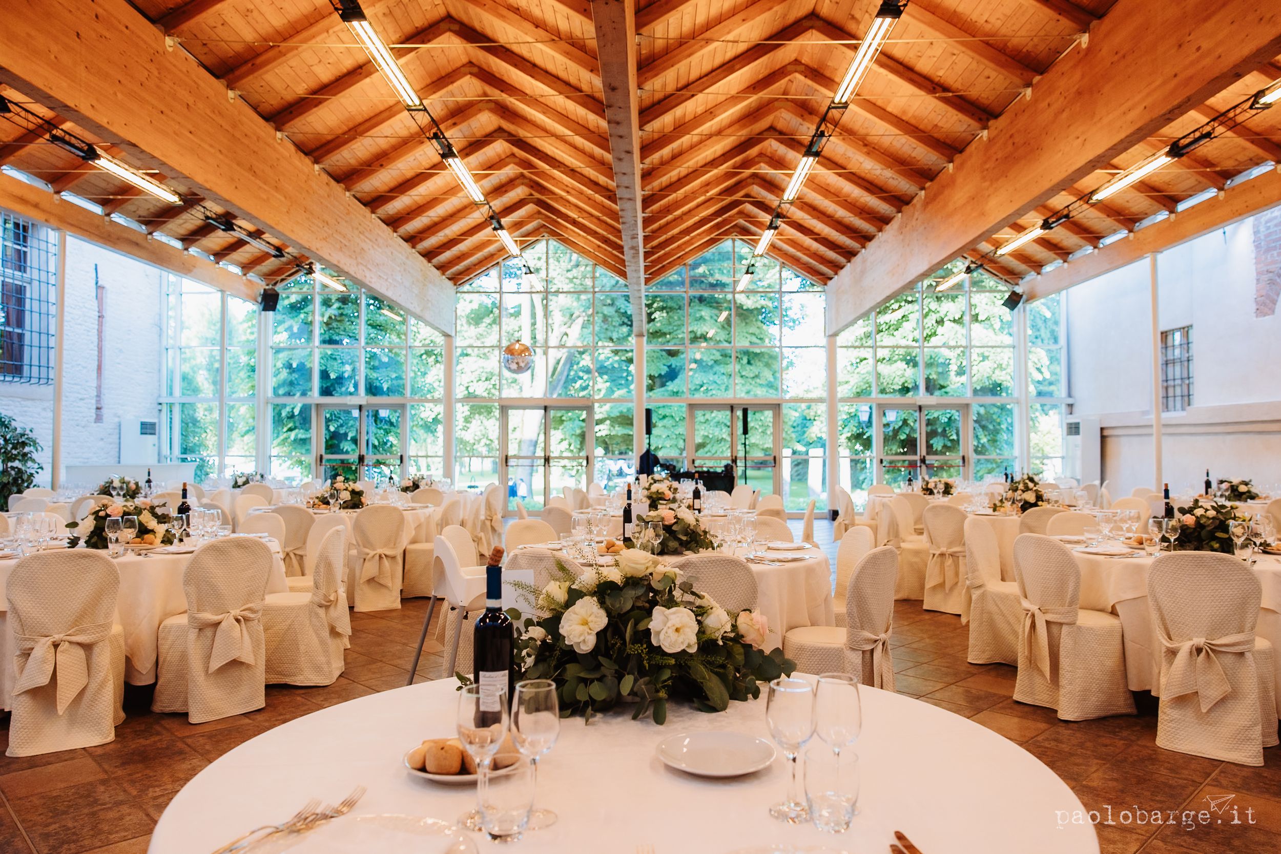 Elegant wedding reception venue with exposed wooden beams, crystal chandeliers, and white-draped tables set for celebration.