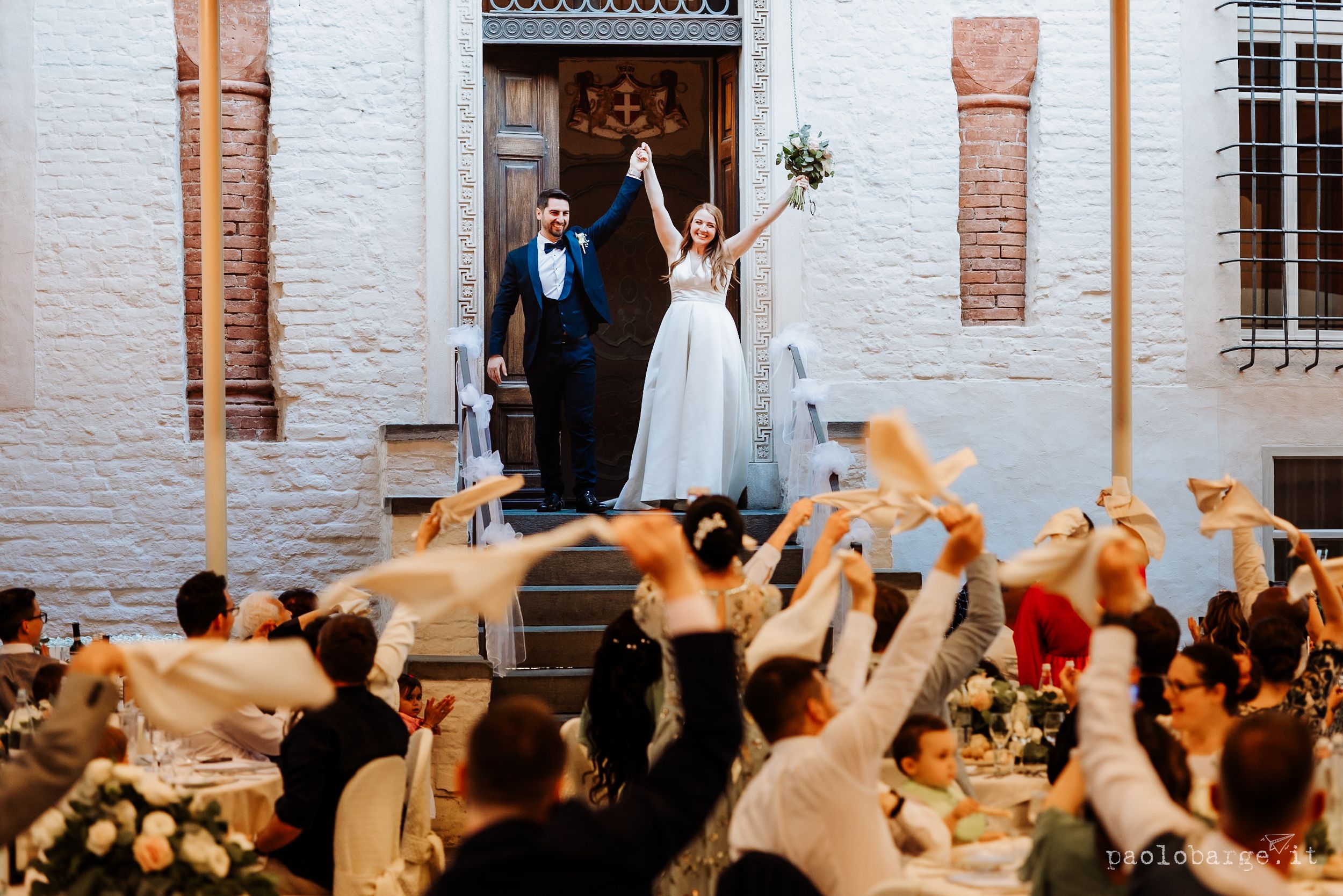 Wedding celebration showing a couple on building steps with an excited crowd raising their hands and phones below.