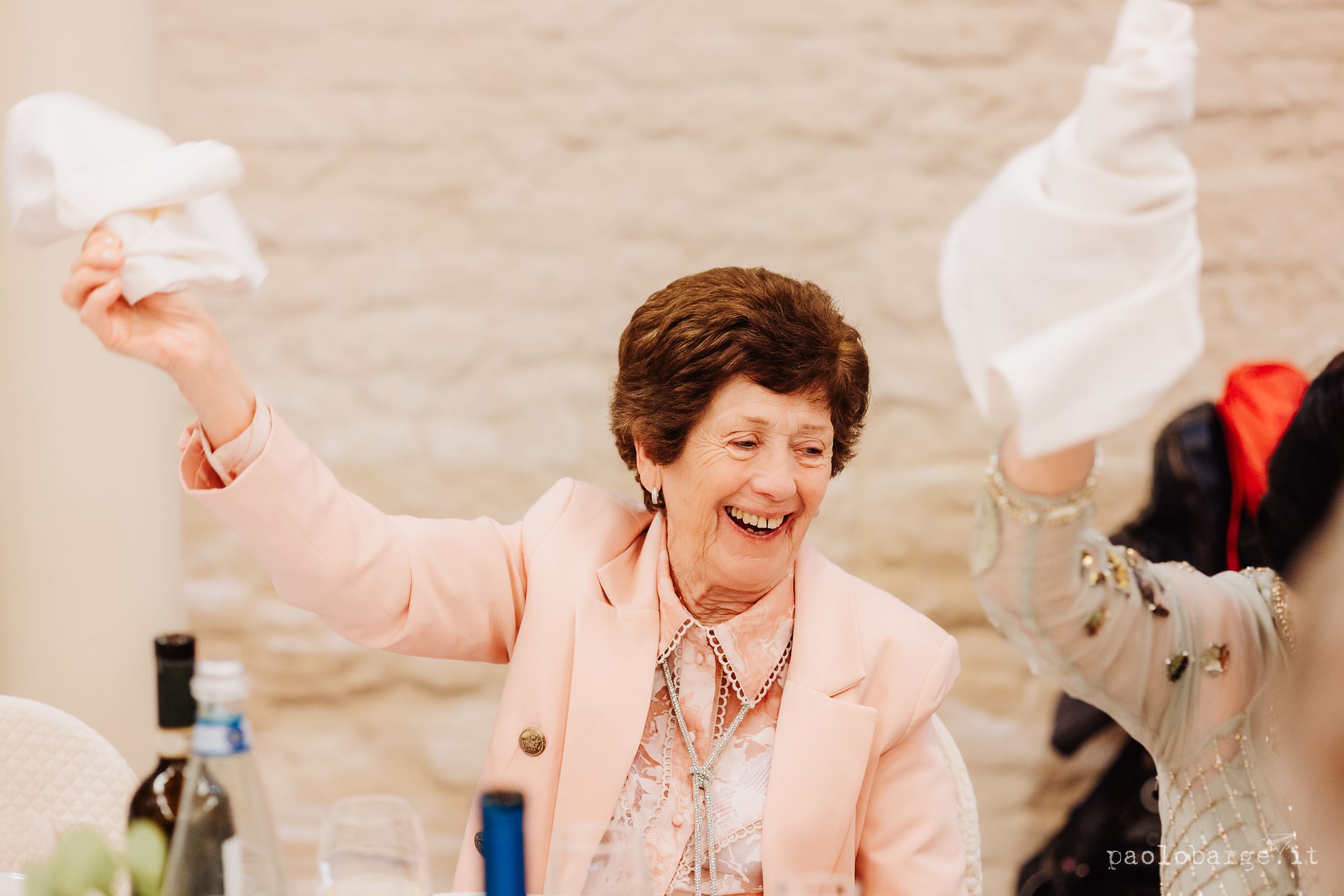 A joyful person in a pink blazer waves napkins while celebrating at a social gathering.