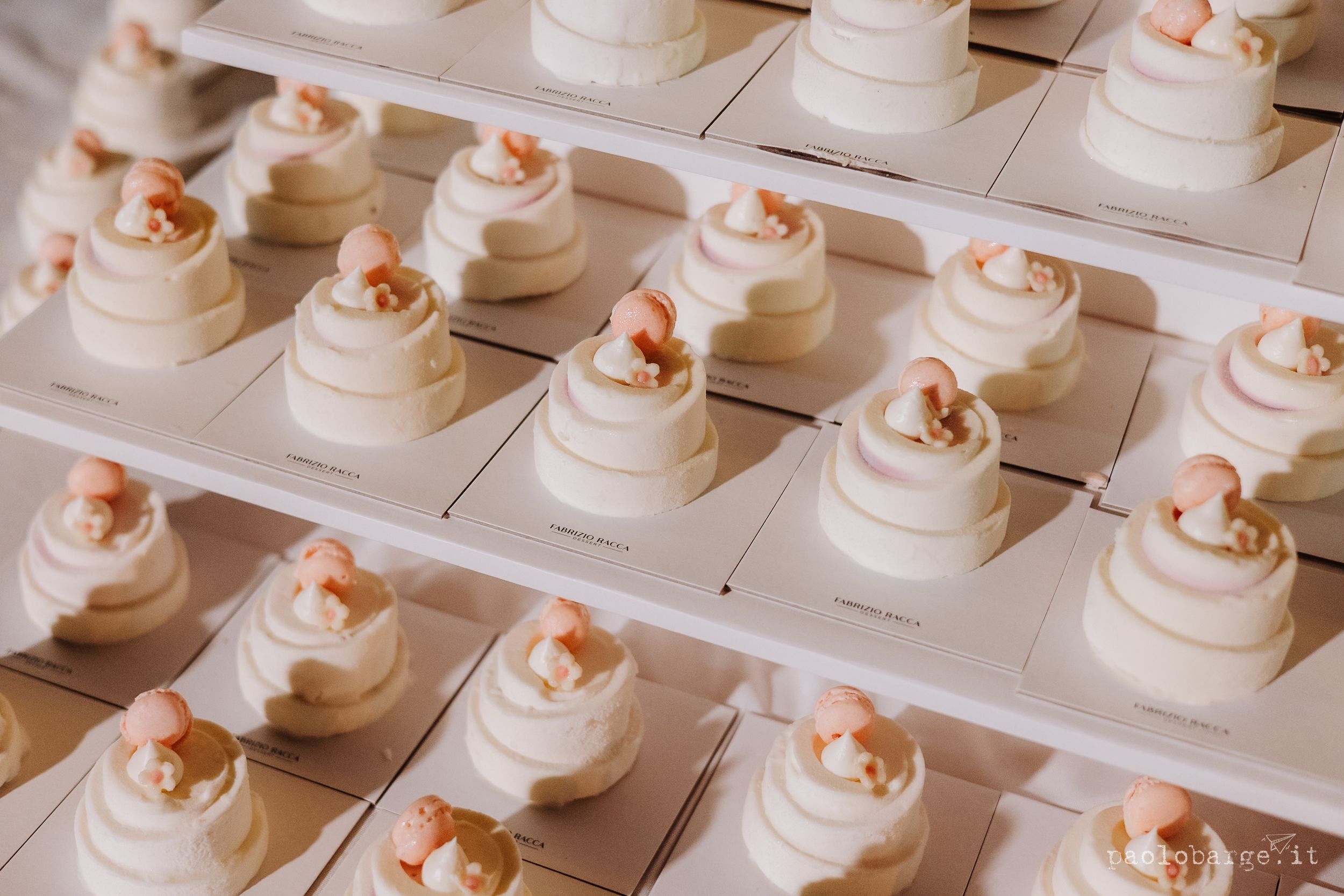 Rows of identical white wedding cupcakes topped with coral-colored frosting flowers arranged in symmetrical pattern on display.