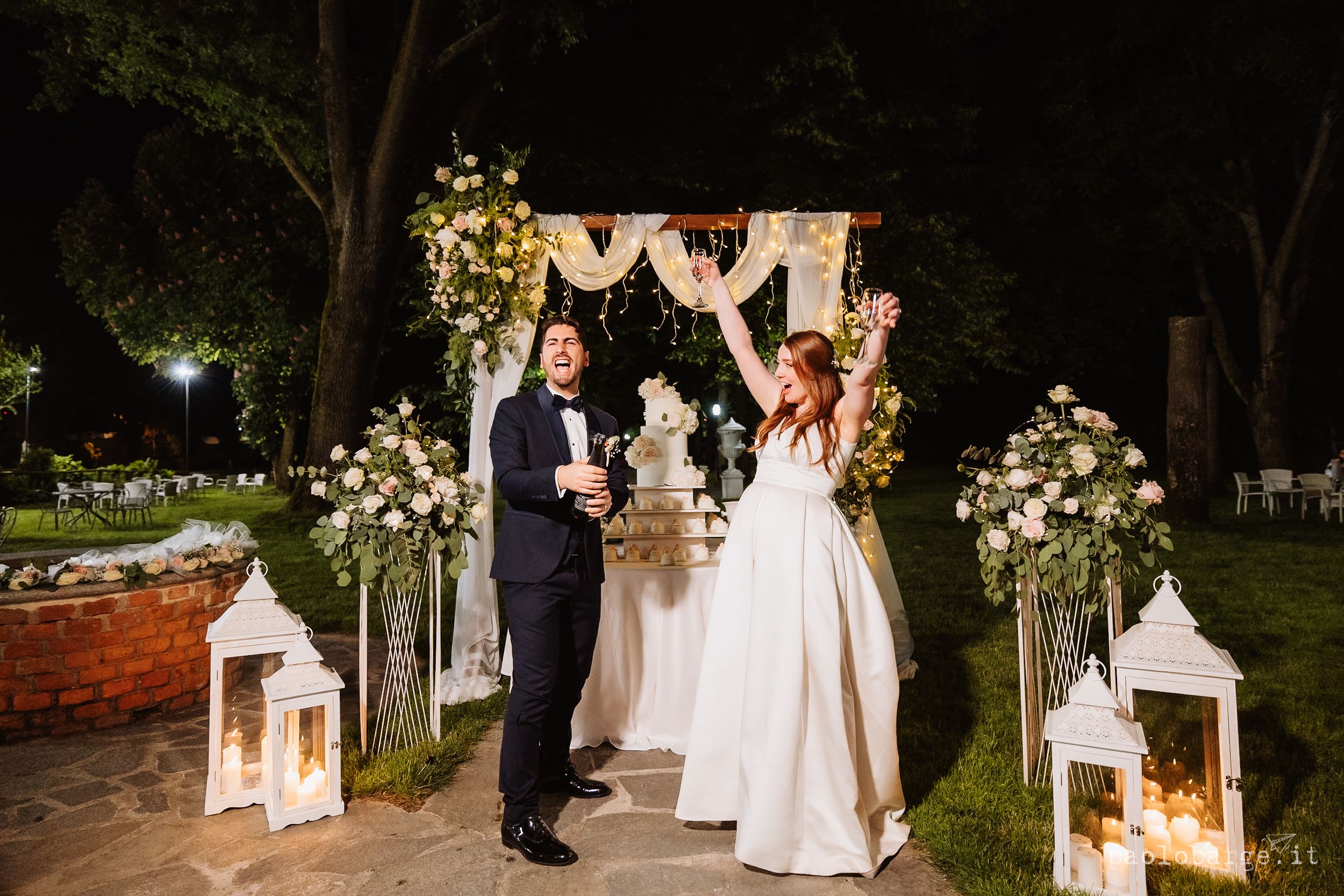 A nighttime outdoor wedding ceremony with illuminated lanterns and floral decorations along a garden path.