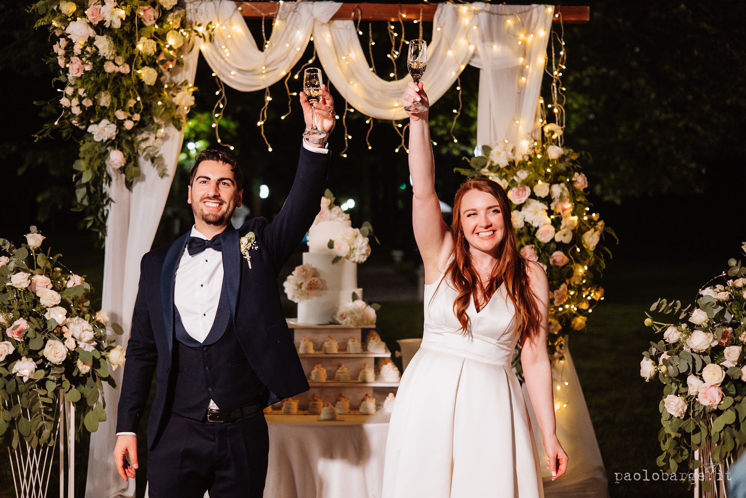 A couple in formal wedding attire shares a joyous moment under an archway decorated with white and yellow flowers.